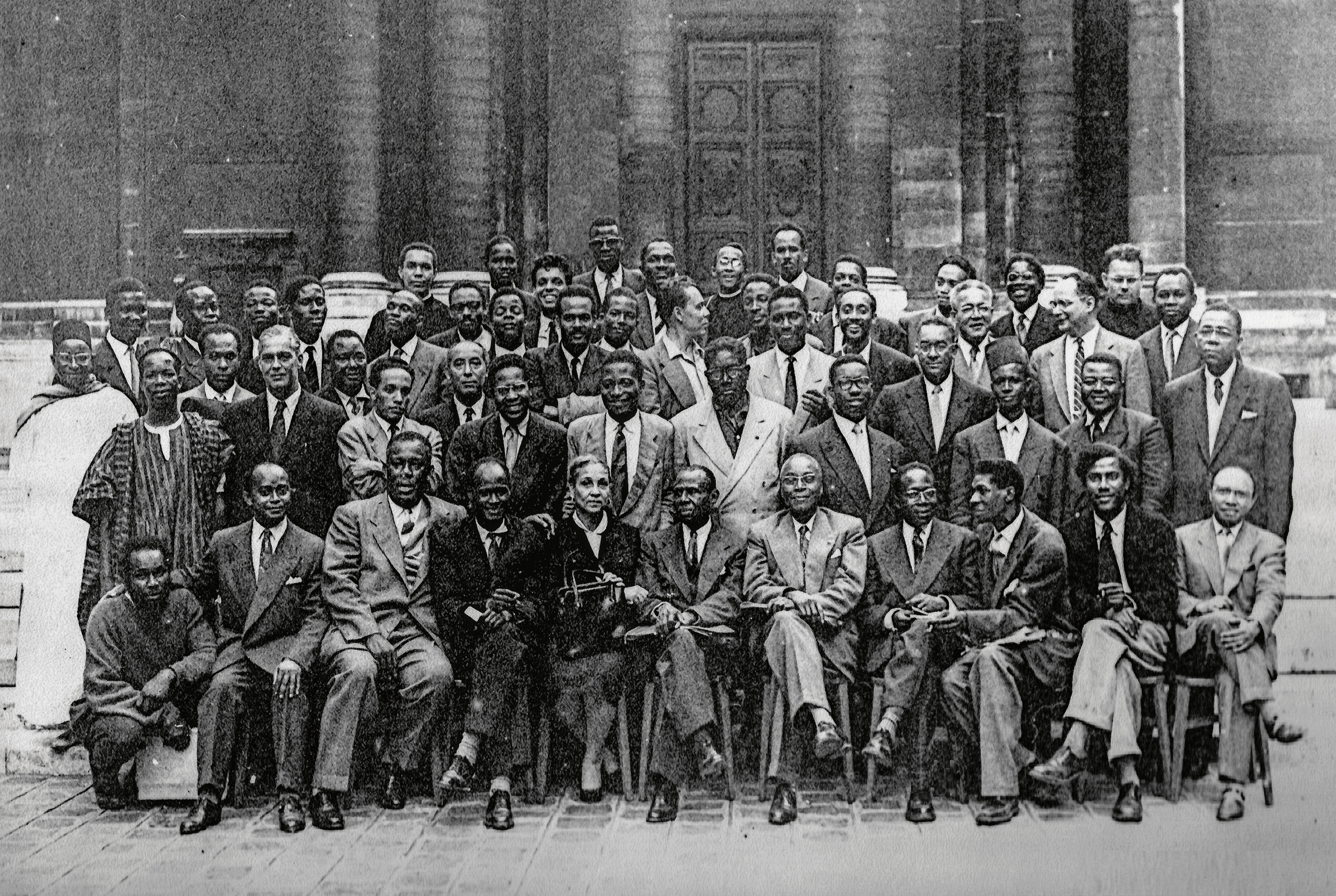 Black and white group portrait of many formally dressed Black men and one woman posing outdoors.
