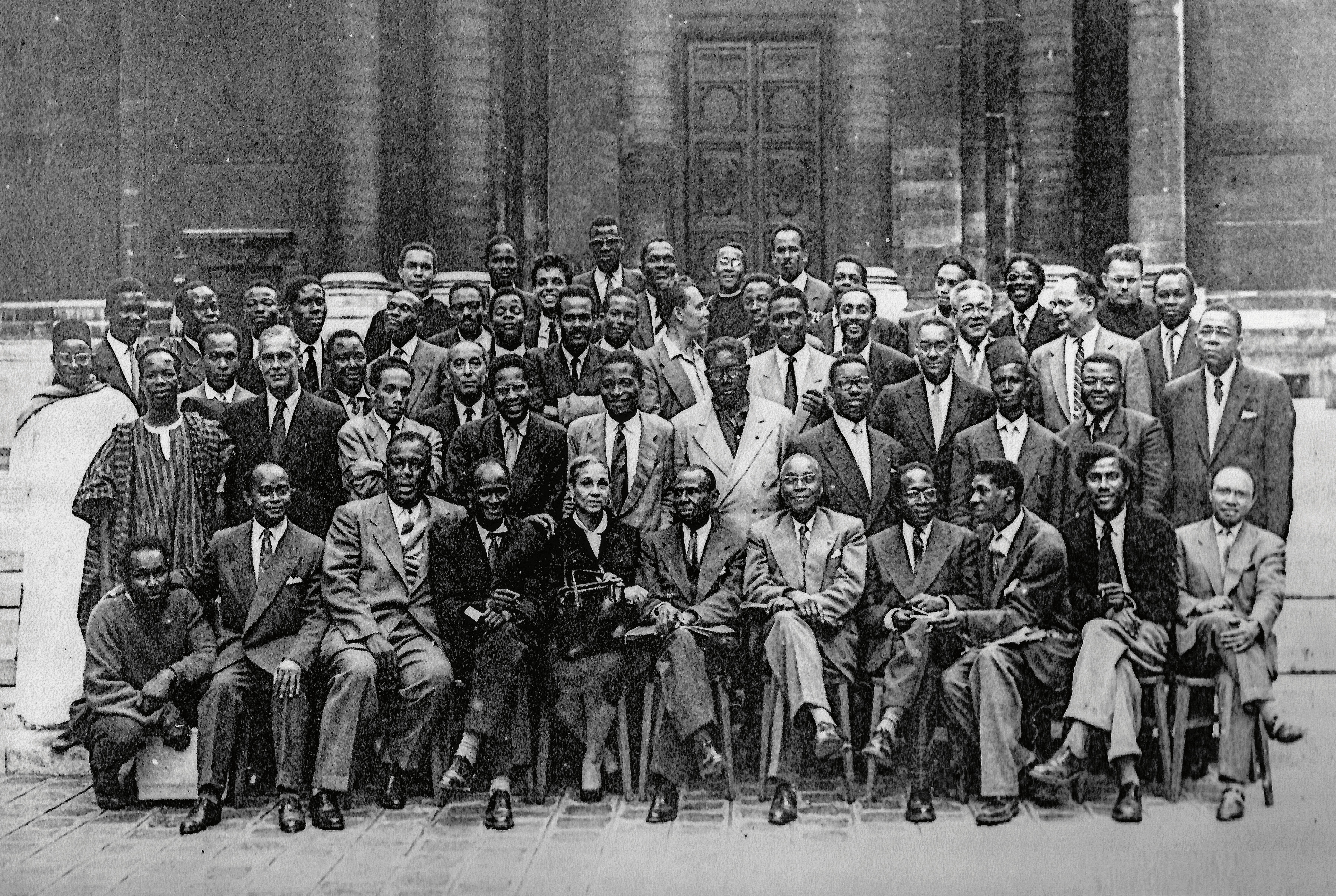 Black and white group portrait of many formally dressed Black men and one woman posing outdoors.