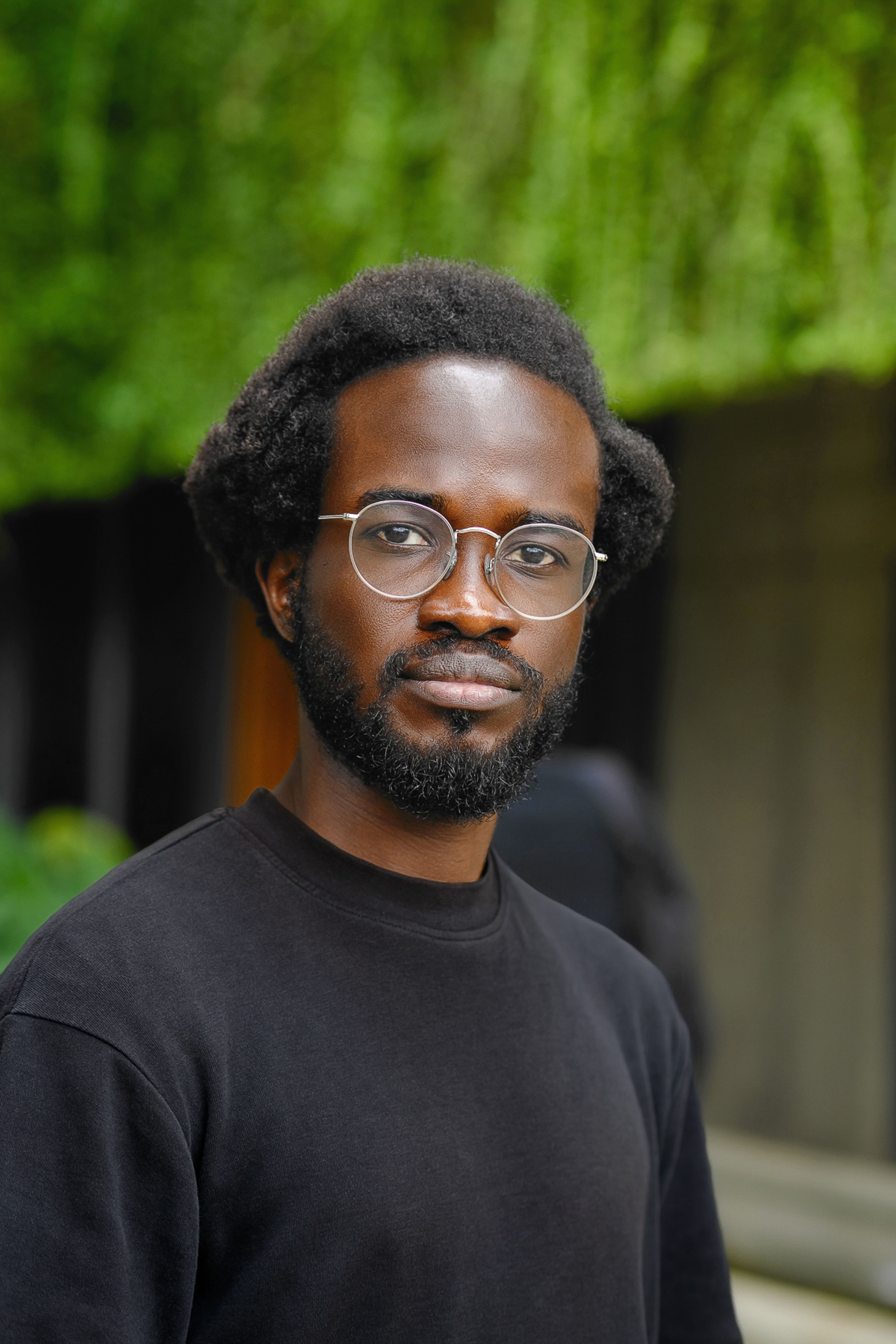 A Black man with short curly hair, a beard, and round glasses wears a black shirt and looks directly at the viewer.