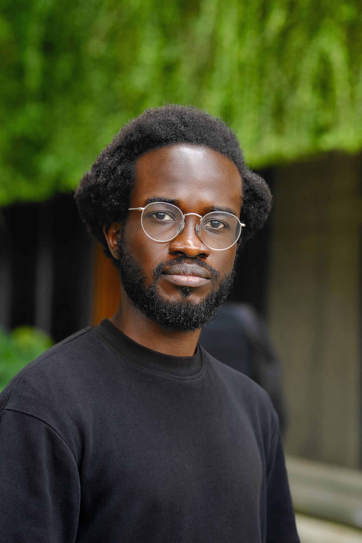 A Black man with short curly hair, a beard, and round glasses wears a black shirt and looks directly at the viewer.