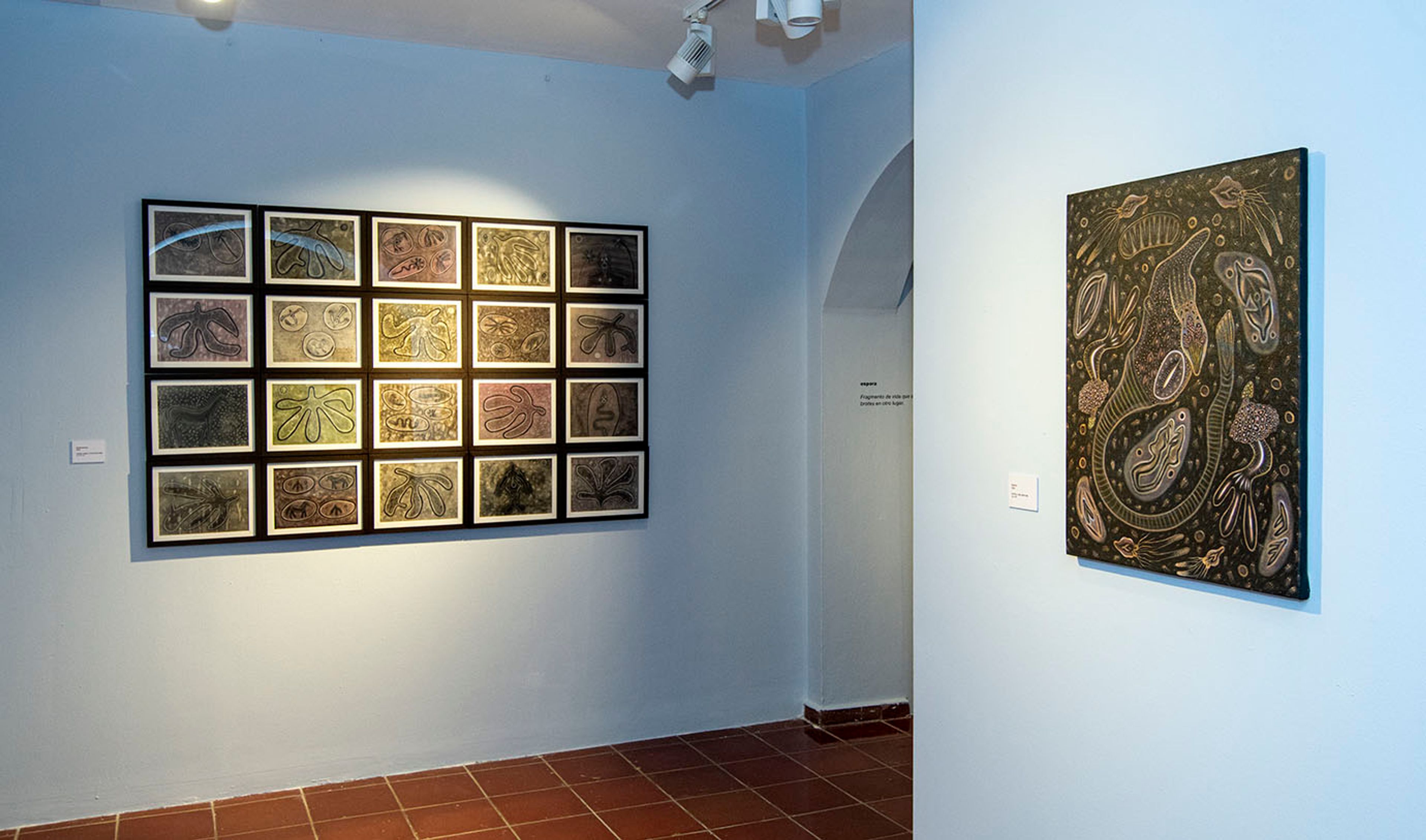 An art gallery with light blue walls, featuring a large grid of 20 framed artworks depicting stylized leaves, and a vertical painting with abstract gold and green organic shapes.