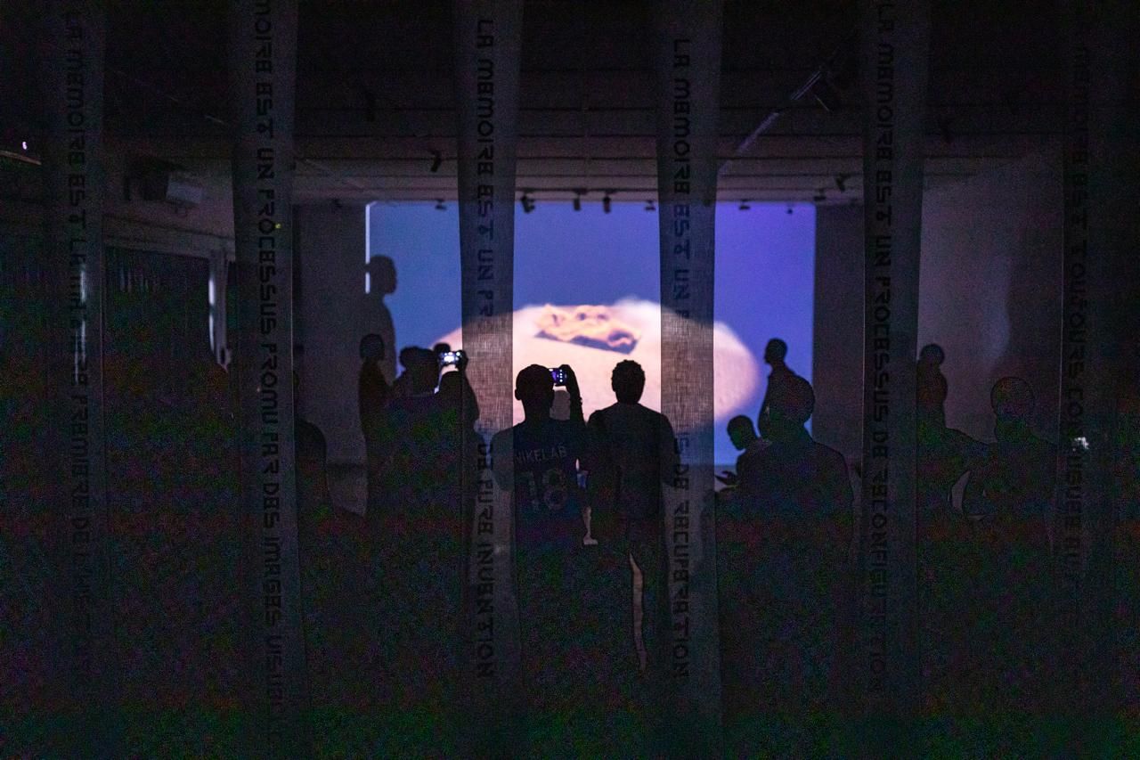 Silhouetted people watch a large screen with a sandy landscape in a dark room, framed by pillars inscribed with French text.