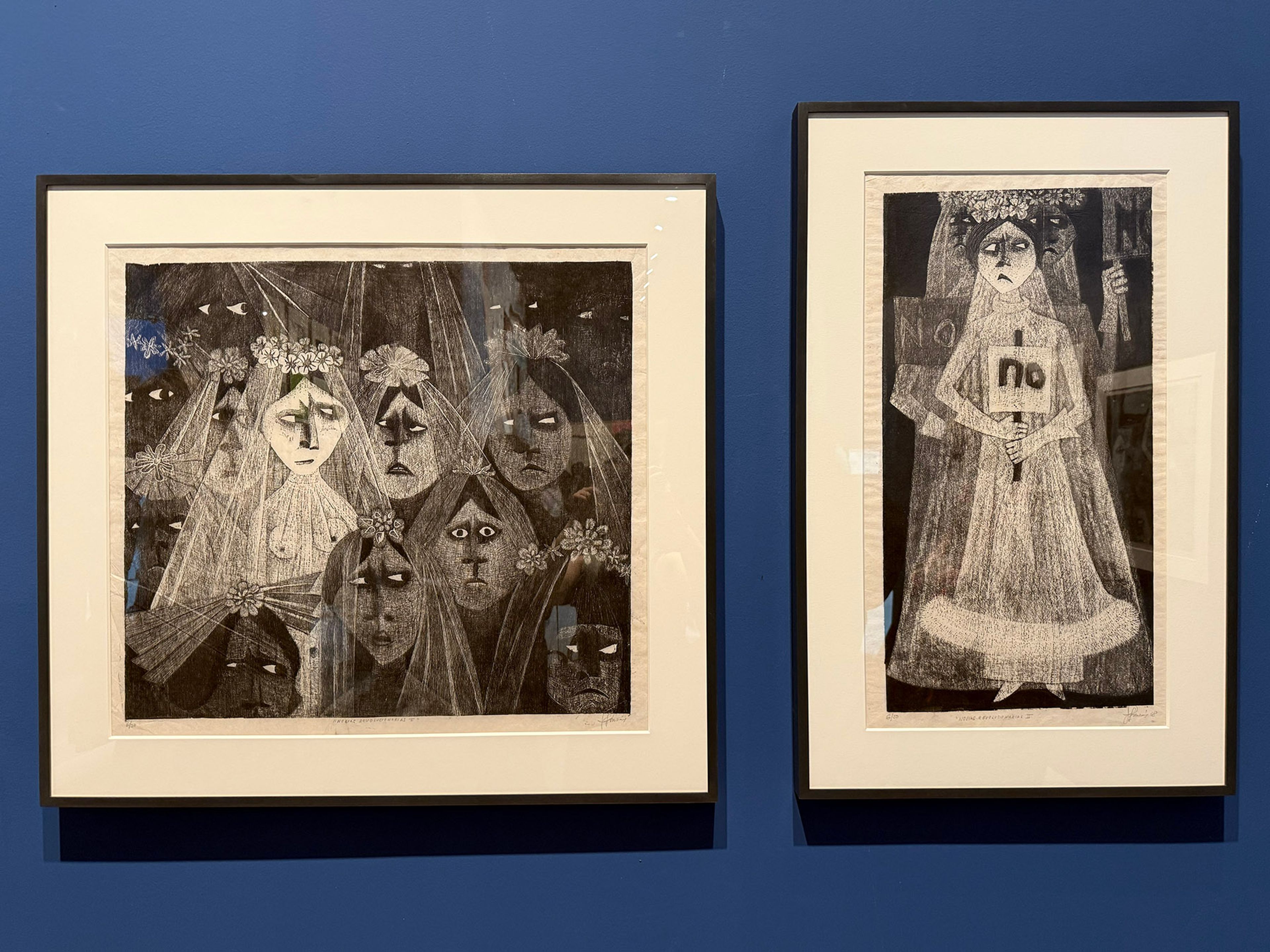 Two framed black and white prints on a blue wall, featuring somber veiled women, with one figure holding a sign that reads 'no'.