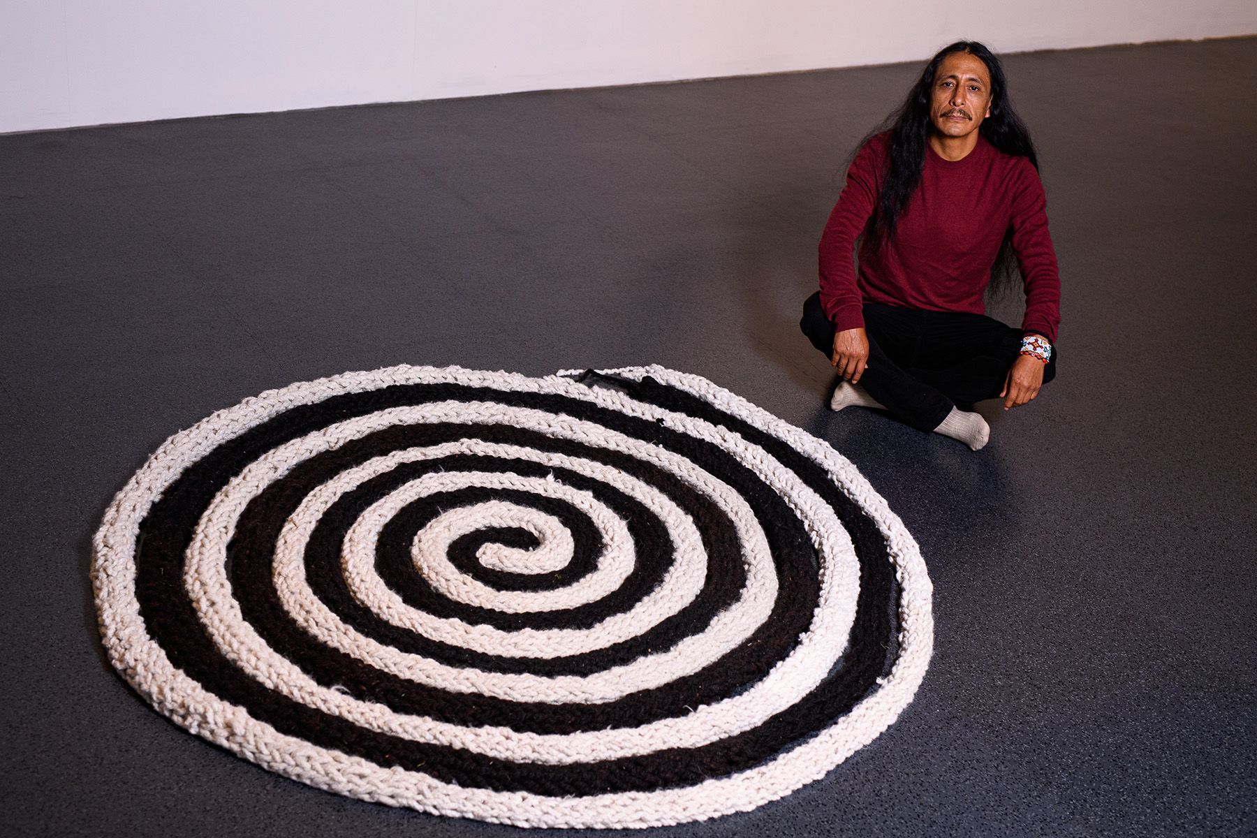 A man with long hair crouches next to a large black and white spiral textile artwork.