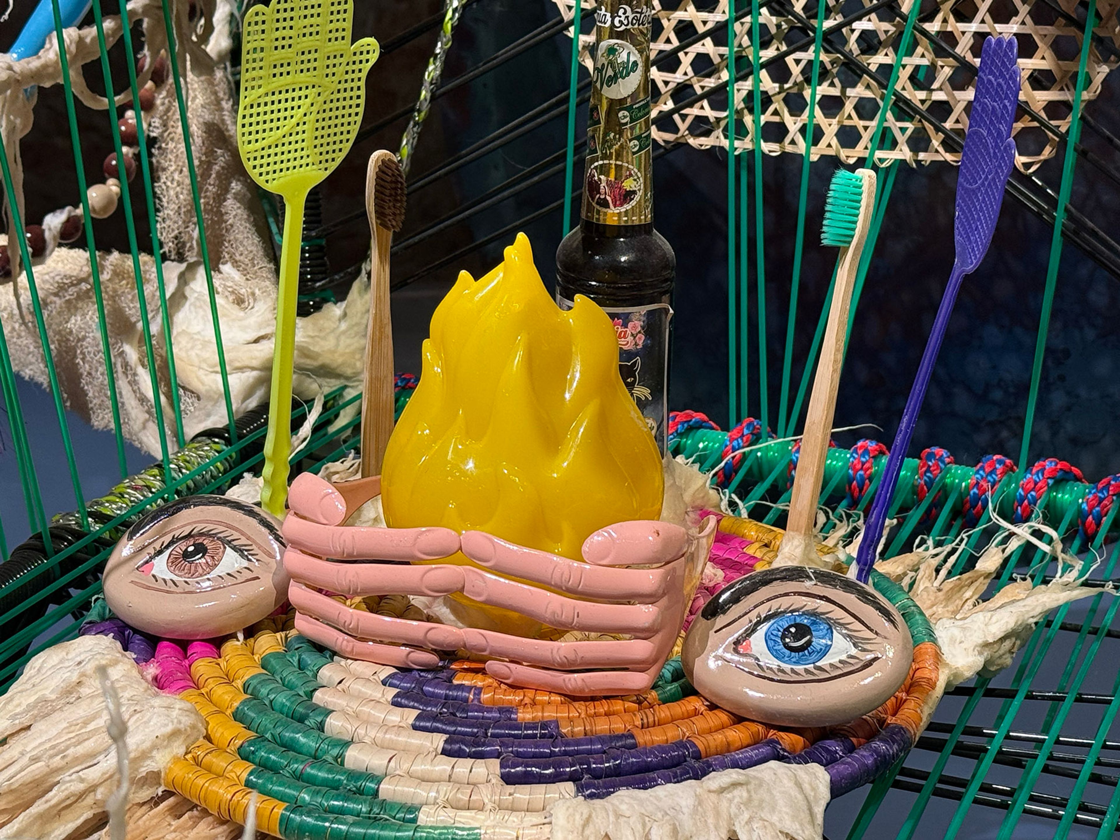 A vibrant arrangement on a woven mat with a yellow flame held by pink hands, two painted eye-rocks, and everyday items like toothbrushes and fly swatters.