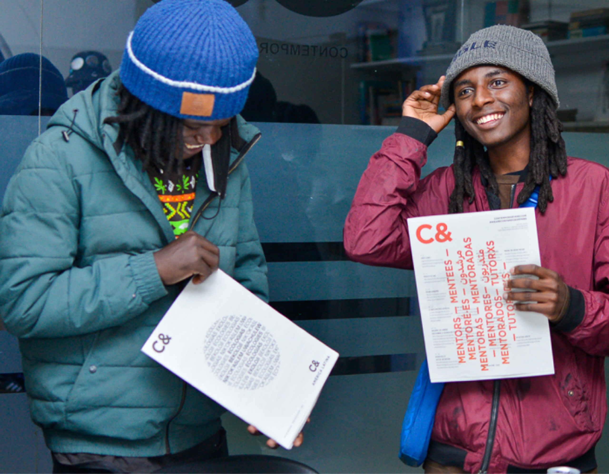 Two young men with dreadlocks, both holding white documents with "C&" branding. The man on the right, smiling, holds up a flyer listing "MENTORS" and "MENTEES."