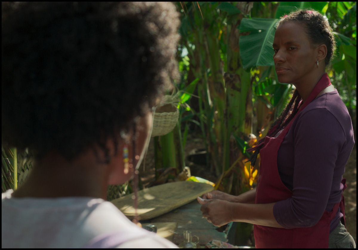 Two Black women outdoors amidst green foliage. One woman, with her back to the viewer, faces another woman wearing a red apron and a serious expression.