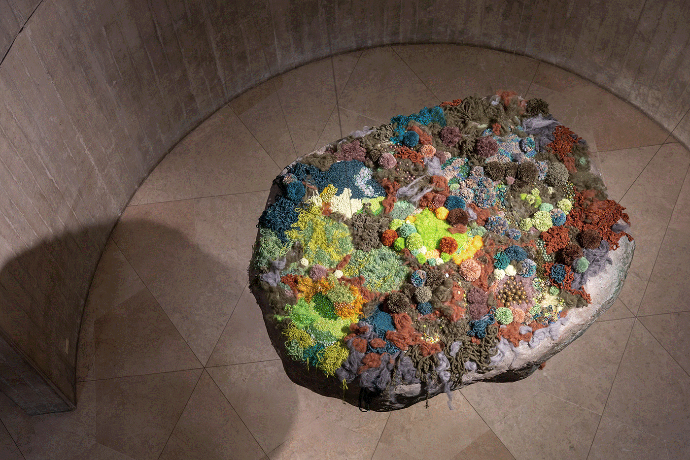 A large, irregular art piece covered in vibrant, multicolored, organic textures, suspended above a light-colored floor.
