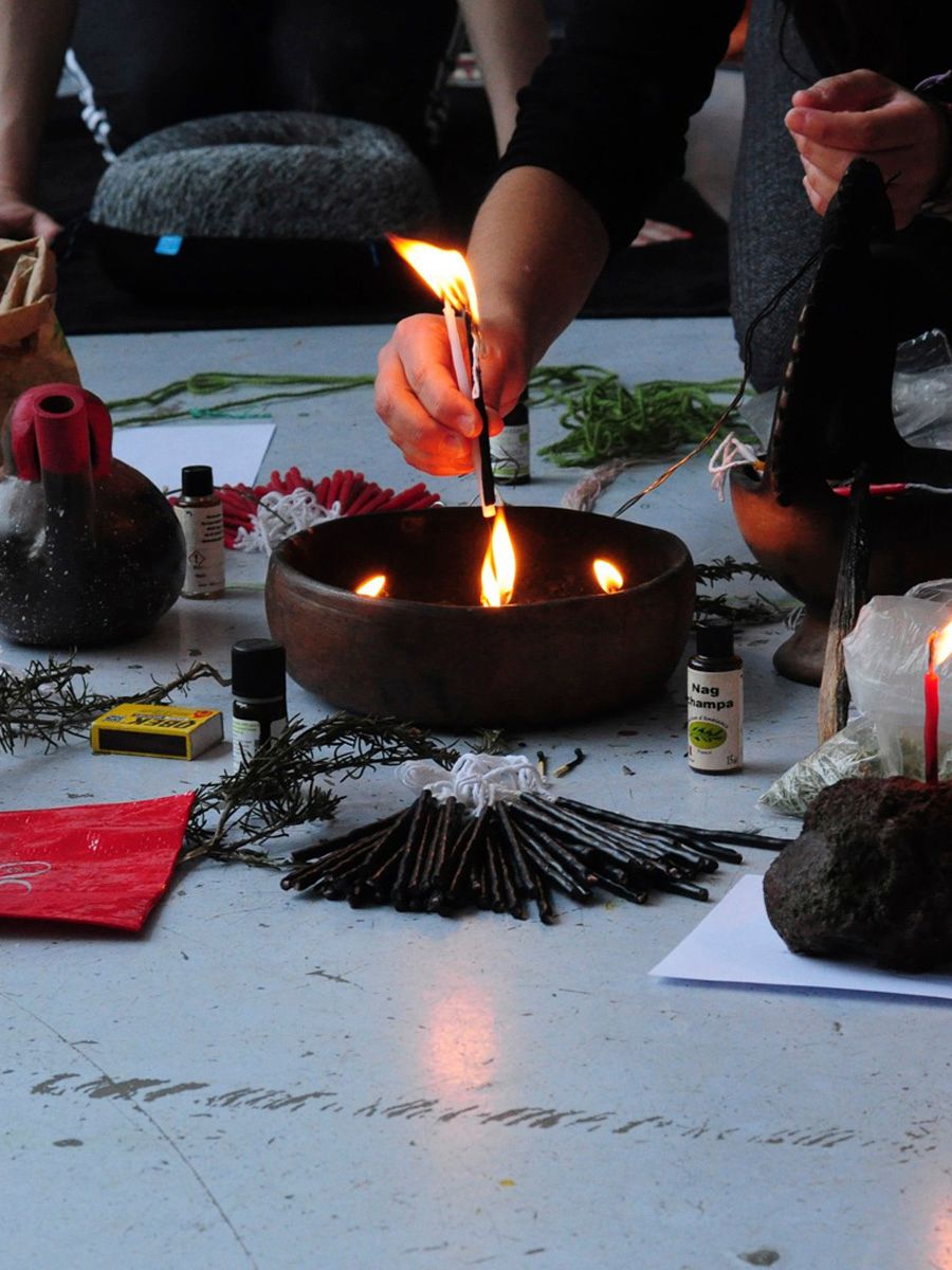 A person's hand lights a small stick from a burning oil lamp in a wooden bowl, surrounded by ritualistic items like herbs and incense sticks.