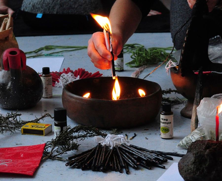 A person's hand lights a small stick from a burning oil lamp in a wooden bowl, surrounded by ritualistic items like herbs and incense sticks.