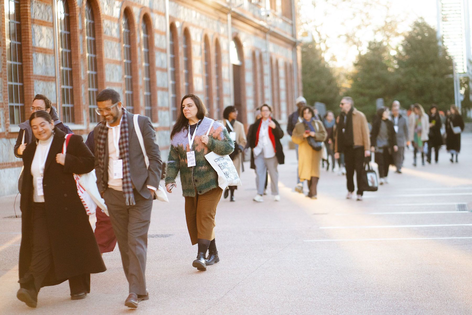 A diverse group of people walks outdoors past a building with arched windows, many wearing event lanyards.