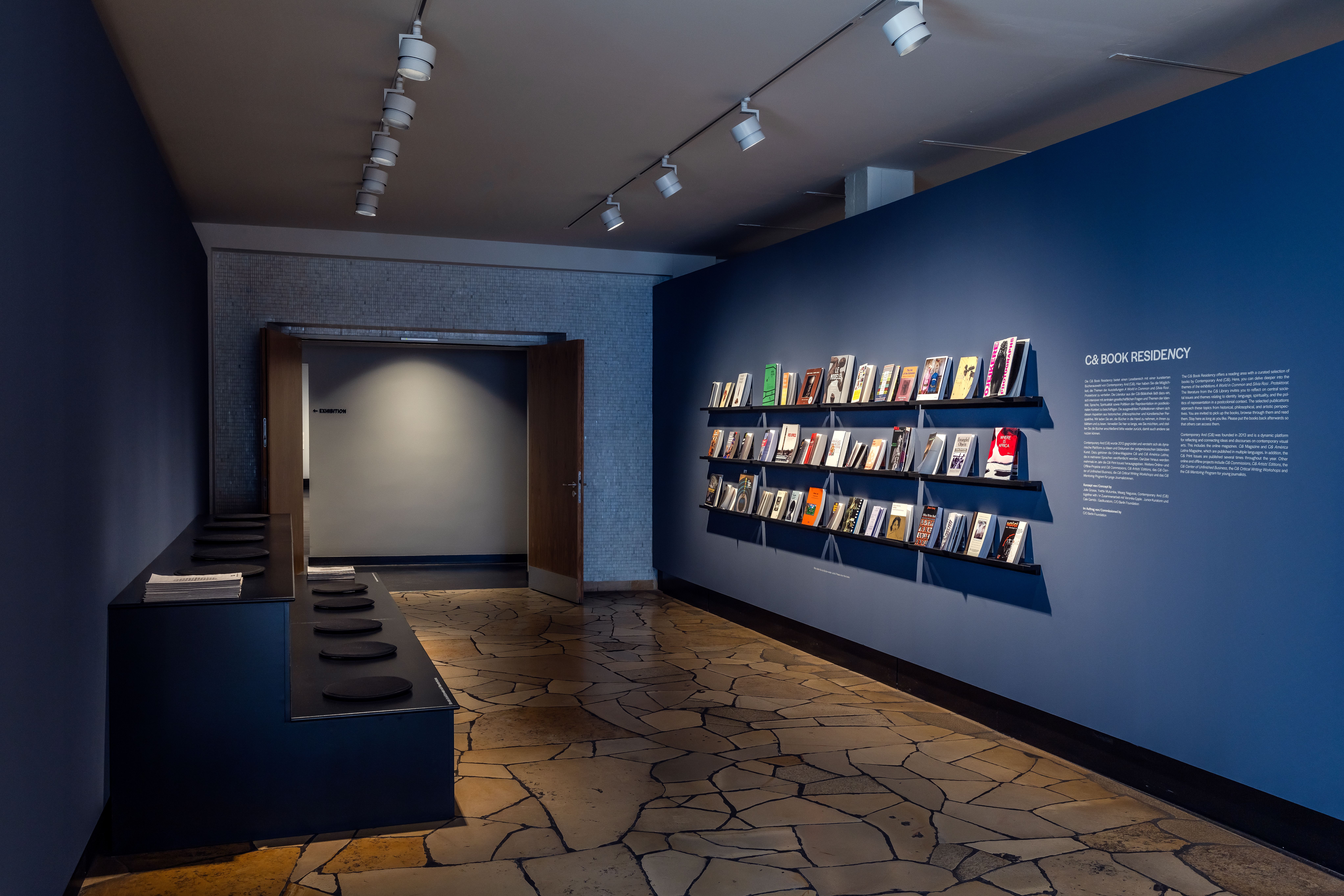 A gallery room with books displayed on shelves on a dark blue wall, accompanied by text for a "CA Book Residency."