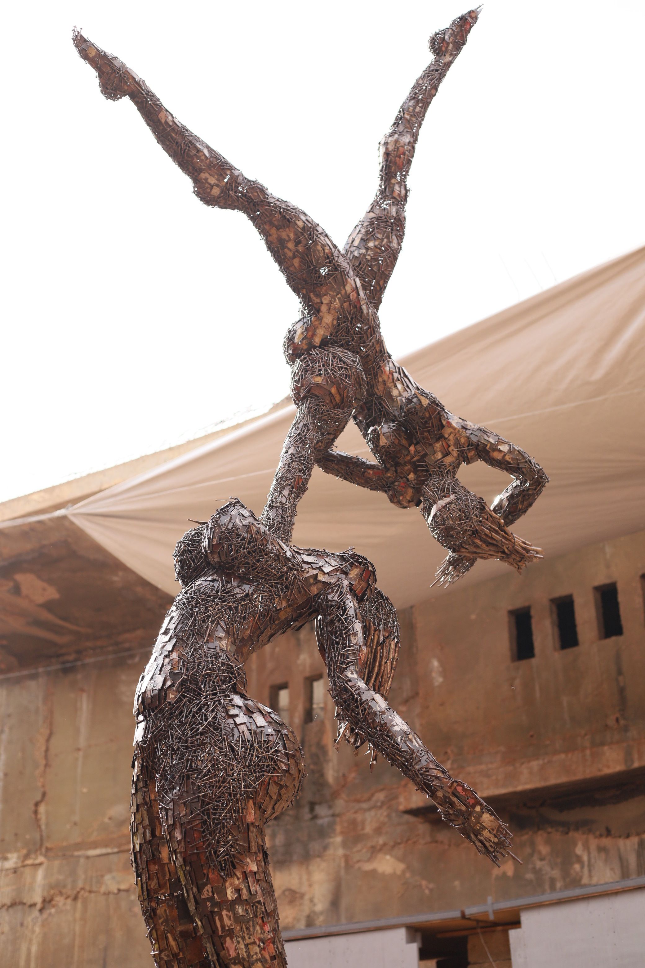 A dynamic sculpture of two human figures crafted from metal fragments, with a pregnant figure balancing an inverted figure on its raised arms.