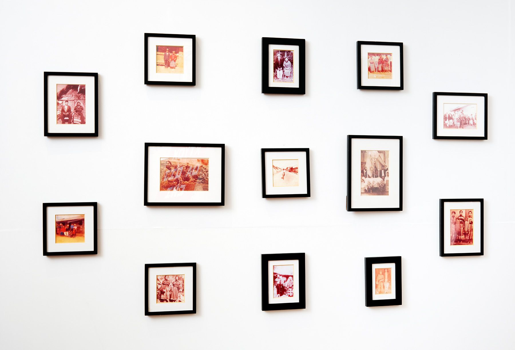 Thirteen framed photos with a reddish-brown hue arranged on a white wall.