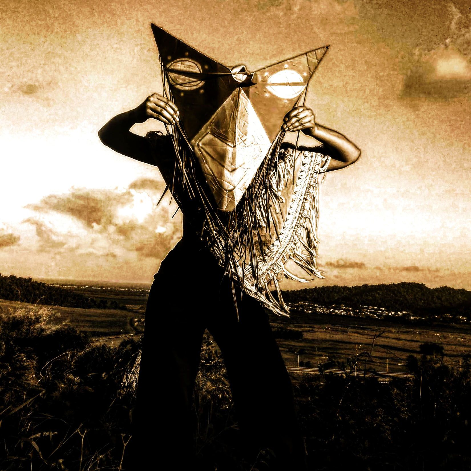 A sepia-toned figure holds a large, star-shaped kite decorated with sun and moon motifs in front of their face, against a dramatic landscape.