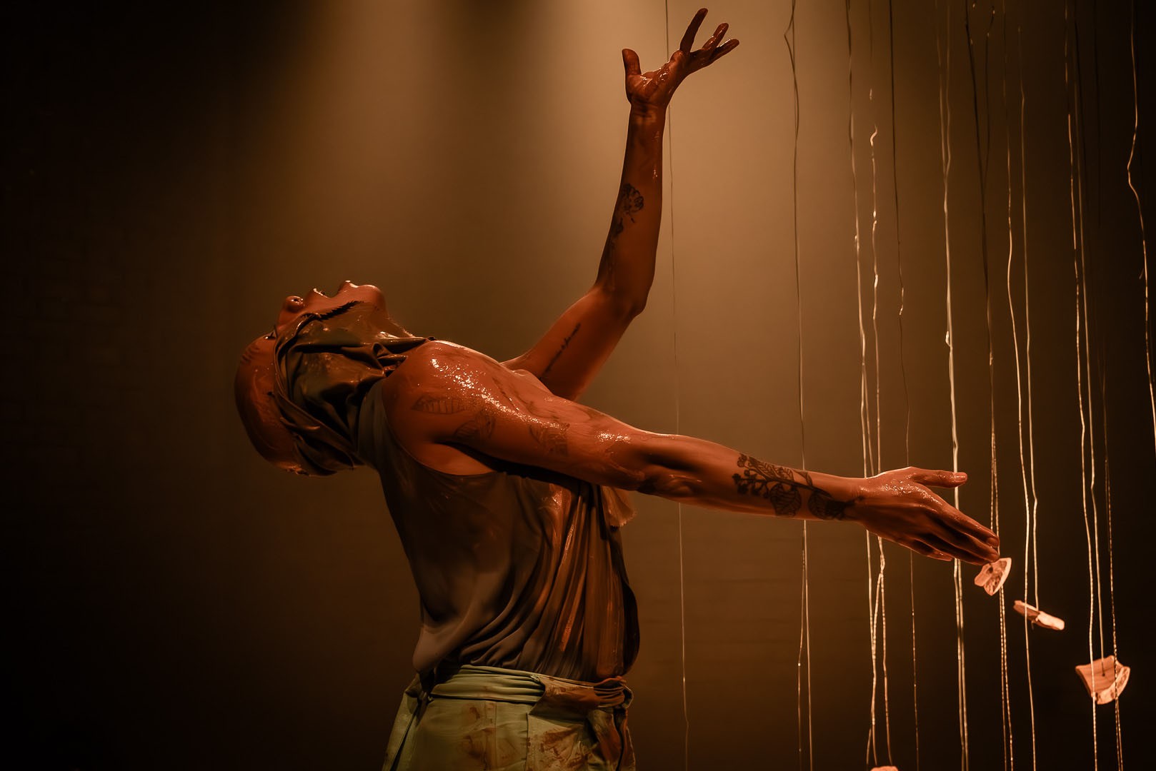 A person drenched in a dark substance, head thrown back and arms raised, under dramatic orange light with falling flakes and thin strands.