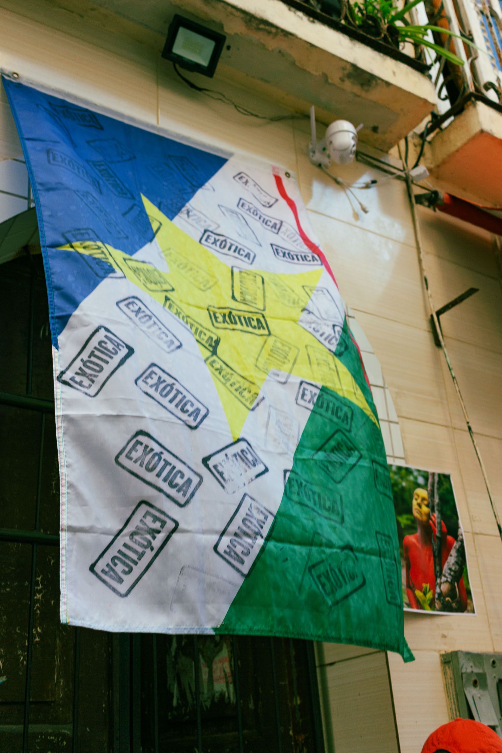 A flag with a yellow star, blue, white, green, and red sections, covered in "EXÓTICA" stamps, hangs on a wall next to a photo of a person in red and yellow paint.