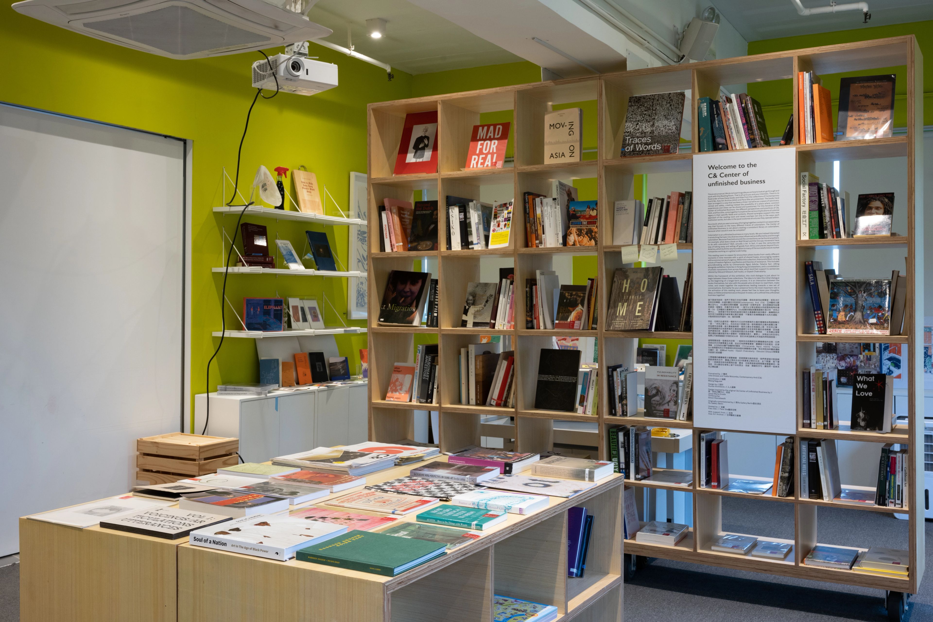 A bright room with lime green walls, wooden bookshelves, and display tables filled with books. A sign on a shelf reads "Welcome to the CB Center of unbounded stillness."