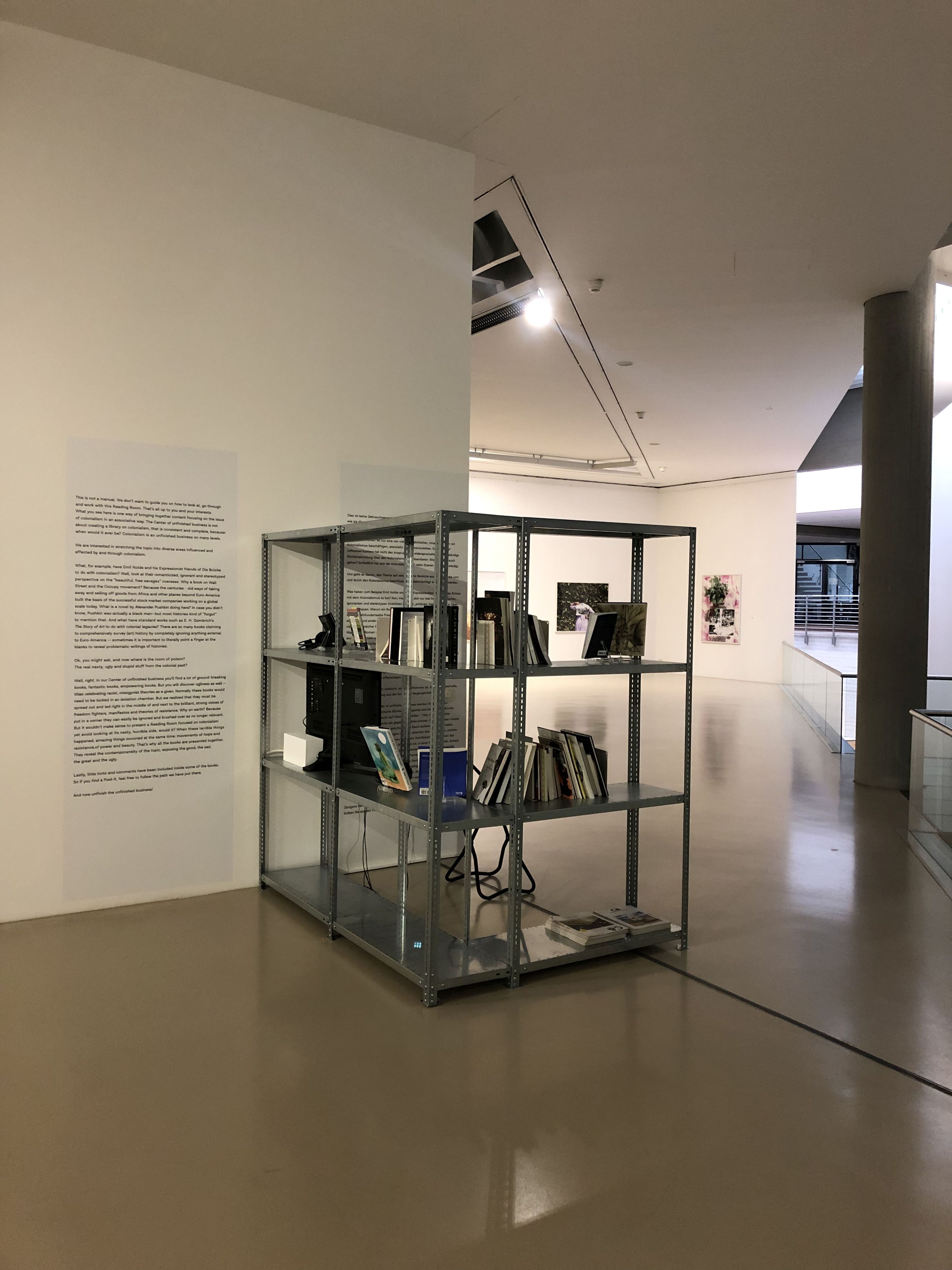 A metal shelving unit filled with books, a monitor, and cables is an art installation in a brightly lit gallery, with wall text on the left and other artworks in the distance.