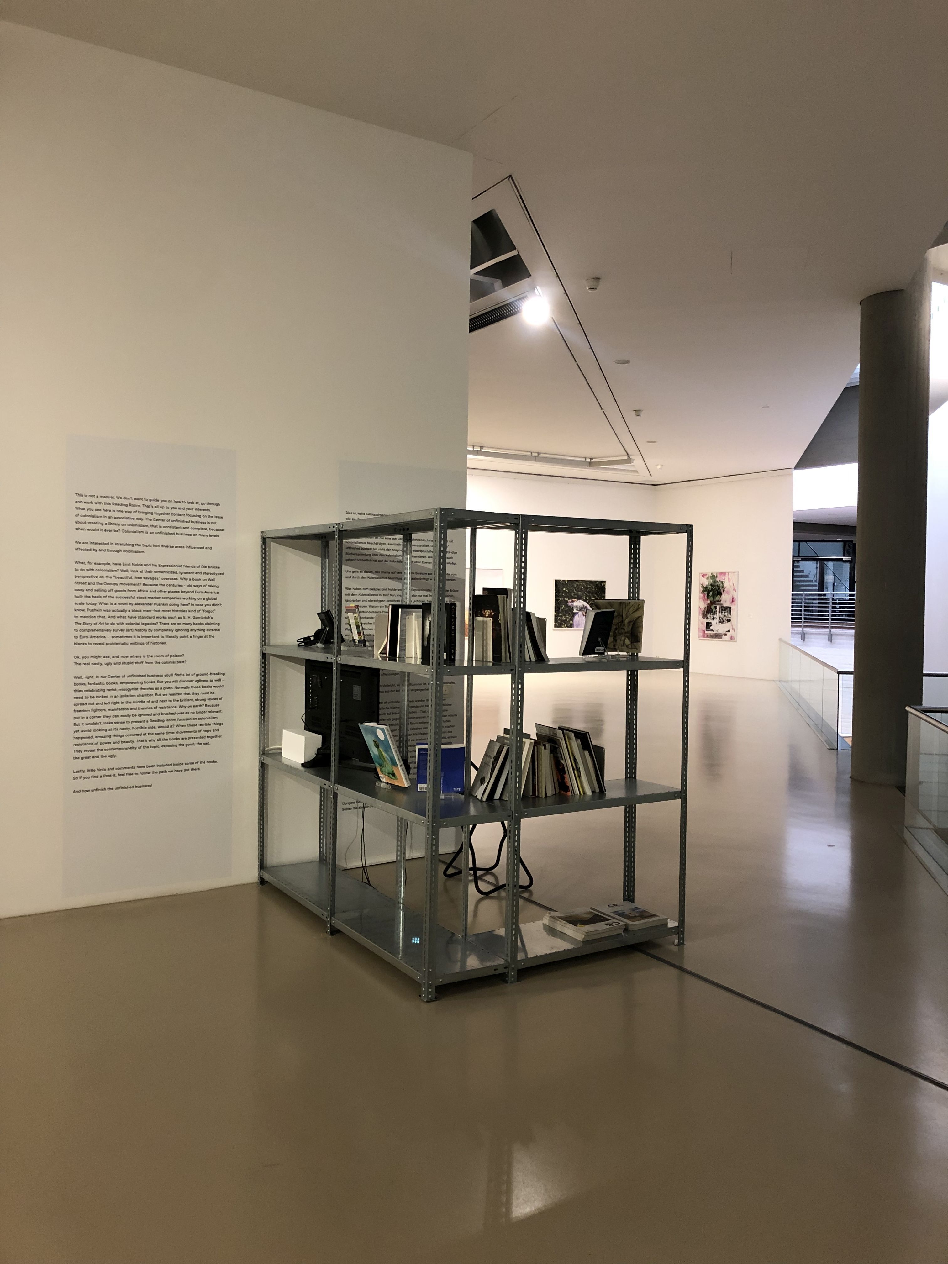A metal shelving unit filled with books, a monitor, and cables is an art installation in a brightly lit gallery, with wall text on the left and other artworks in the distance.