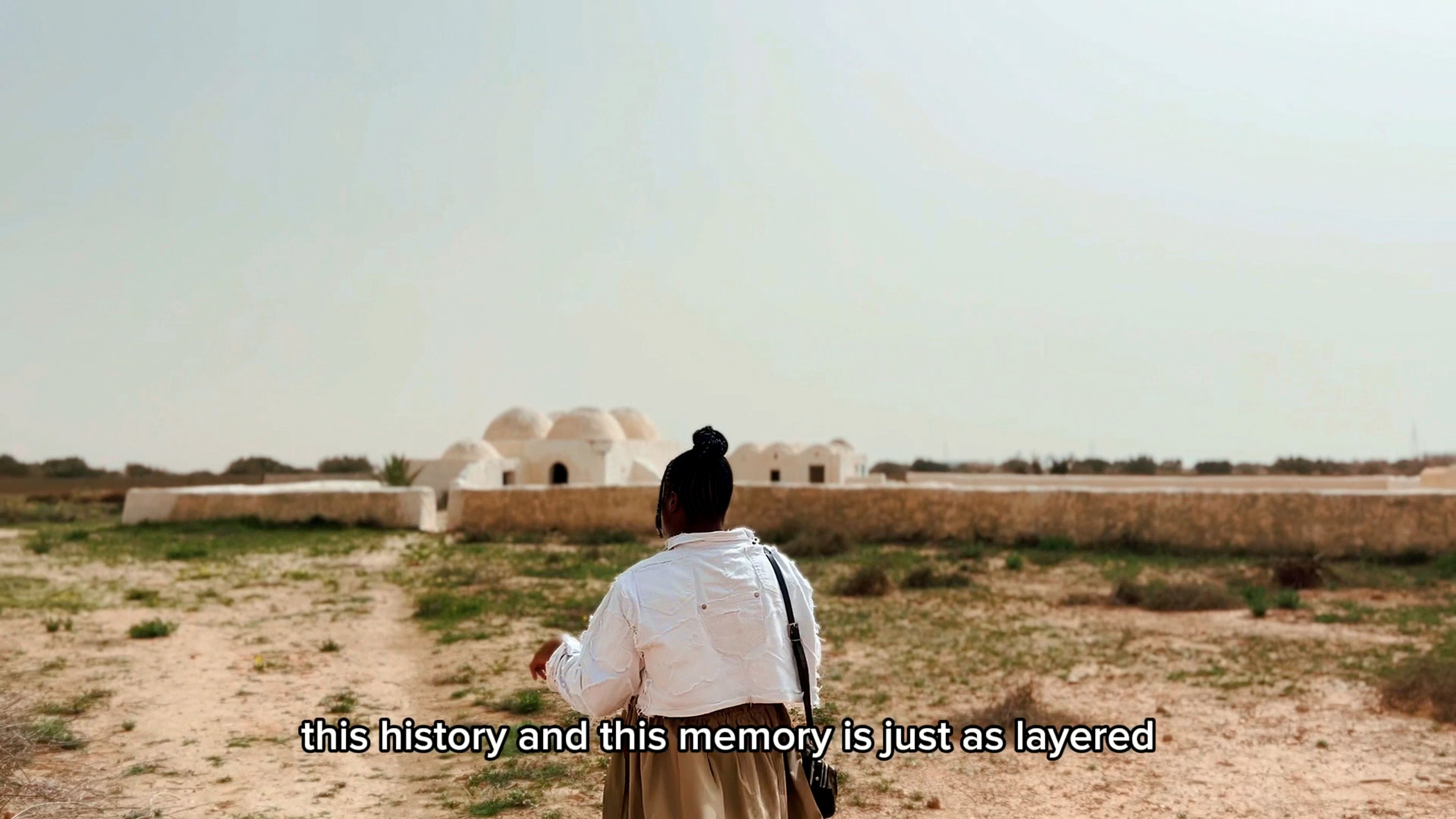 A person facing away stands in a dry, sparse landscape, looking towards white domed buildings in the distance. Subtitles read "this history and this memory is just as layered".