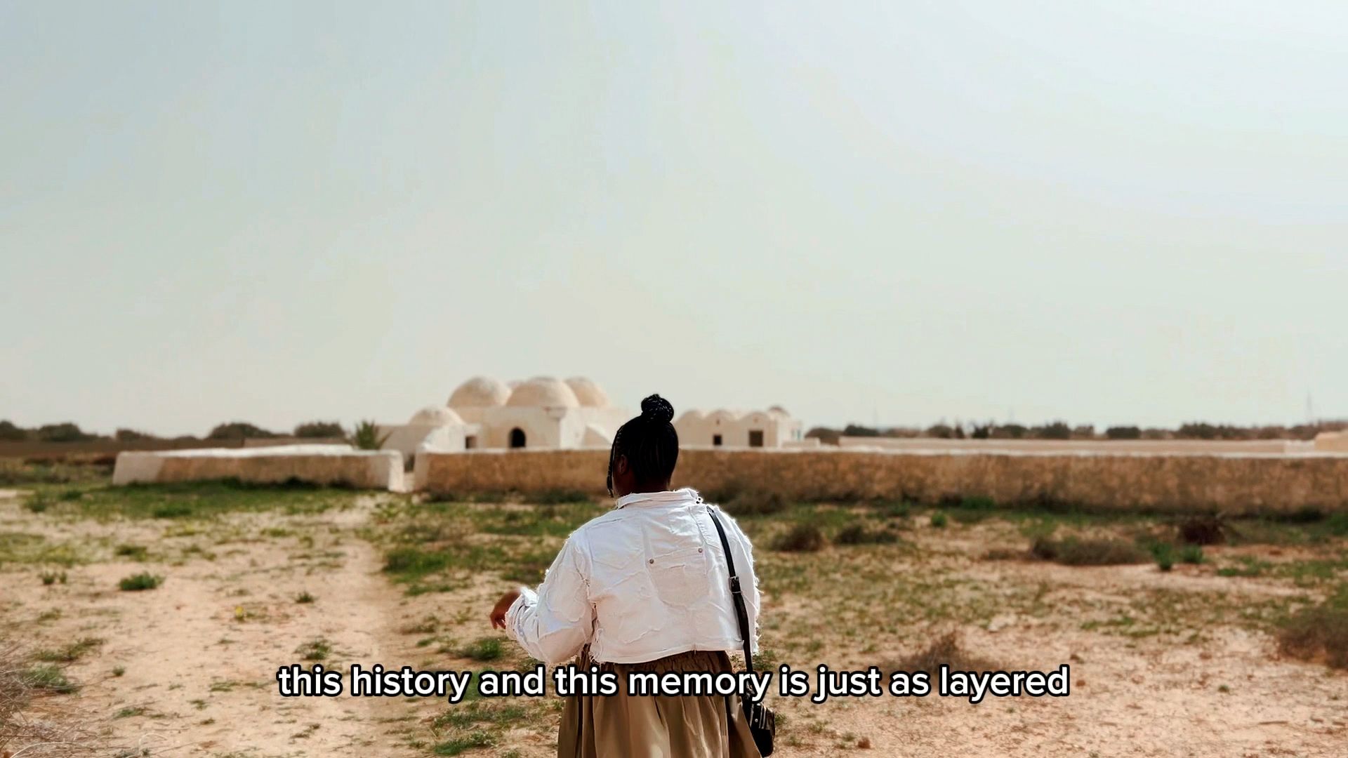 A person facing away stands in a dry, sparse landscape, looking towards white domed buildings in the distance. Subtitles read "this history and this memory is just as layered".