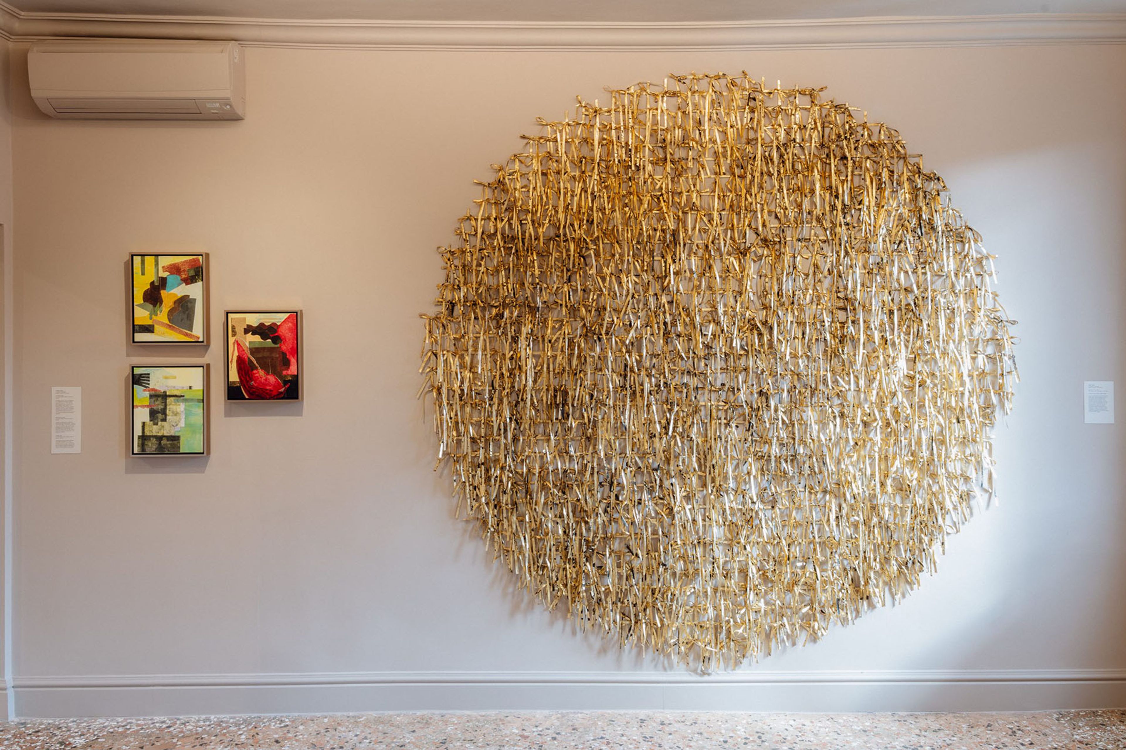 A large, textured golden circular sculpture and three smaller abstract paintings hang on a light gray wall.
