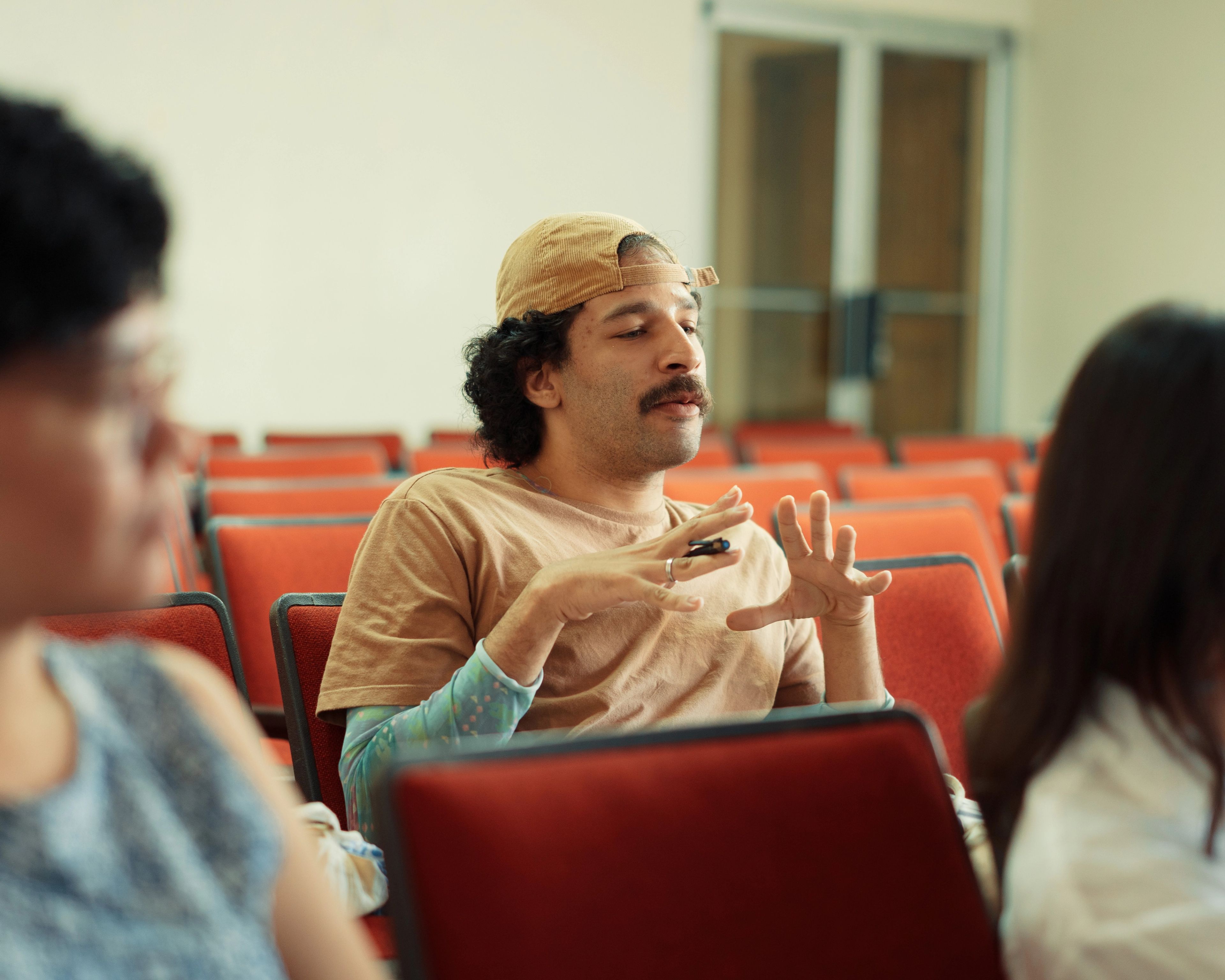 A man in a backward cap and brown t-shirt gestures with his hands while sitting in a lecture hall.