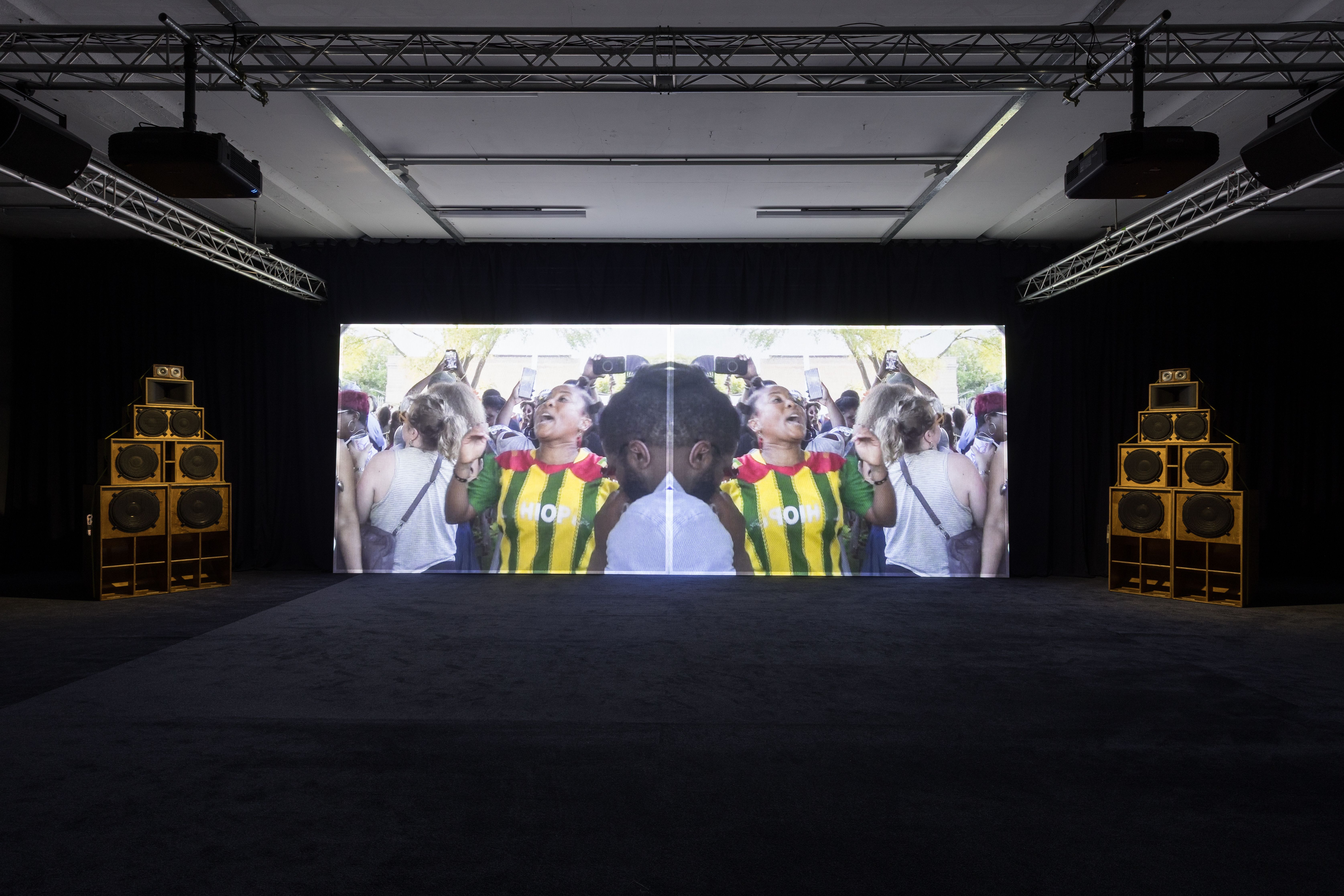 A dark room with a large screen showing a mirrored video of a woman in a striped shirt cheering, flanked by two tall wooden sound system speaker stacks.