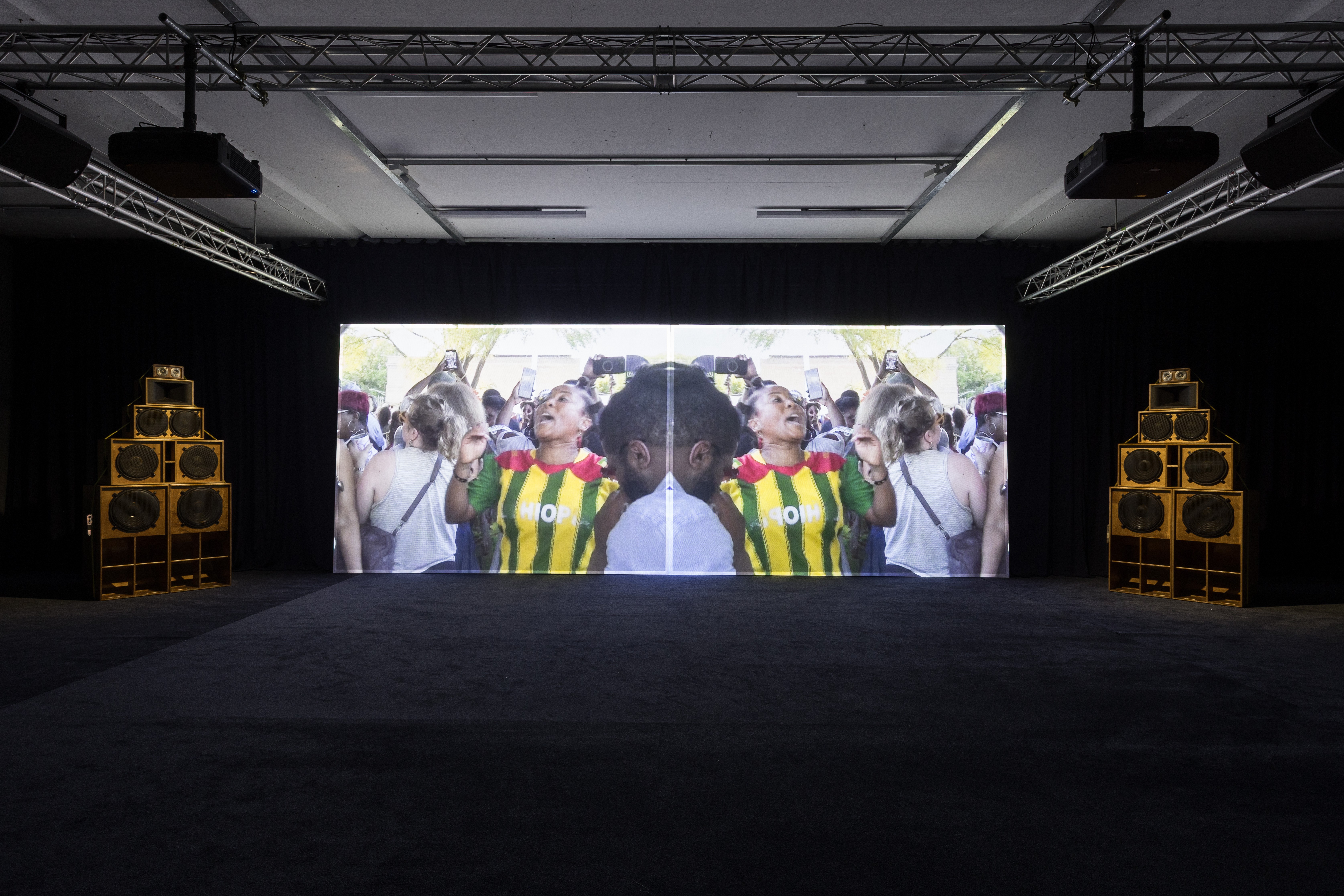 A dark room with a large screen showing a mirrored video of a woman in a striped shirt cheering, flanked by two tall wooden sound system speaker stacks.