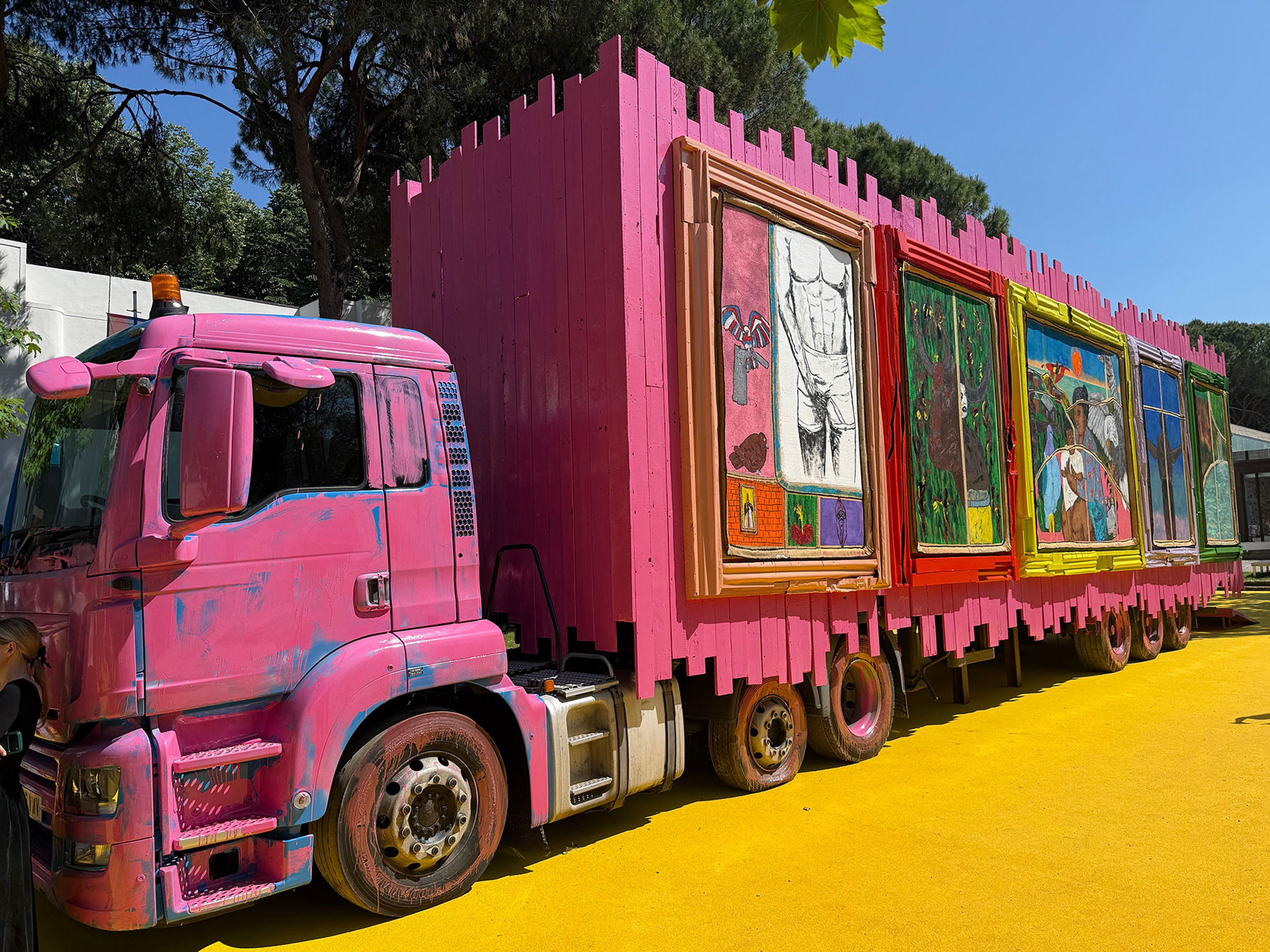 A large pink truck and trailer, with the trailer styled like a castle wall displaying framed artworks, rests on vibrant yellow ground outdoors.