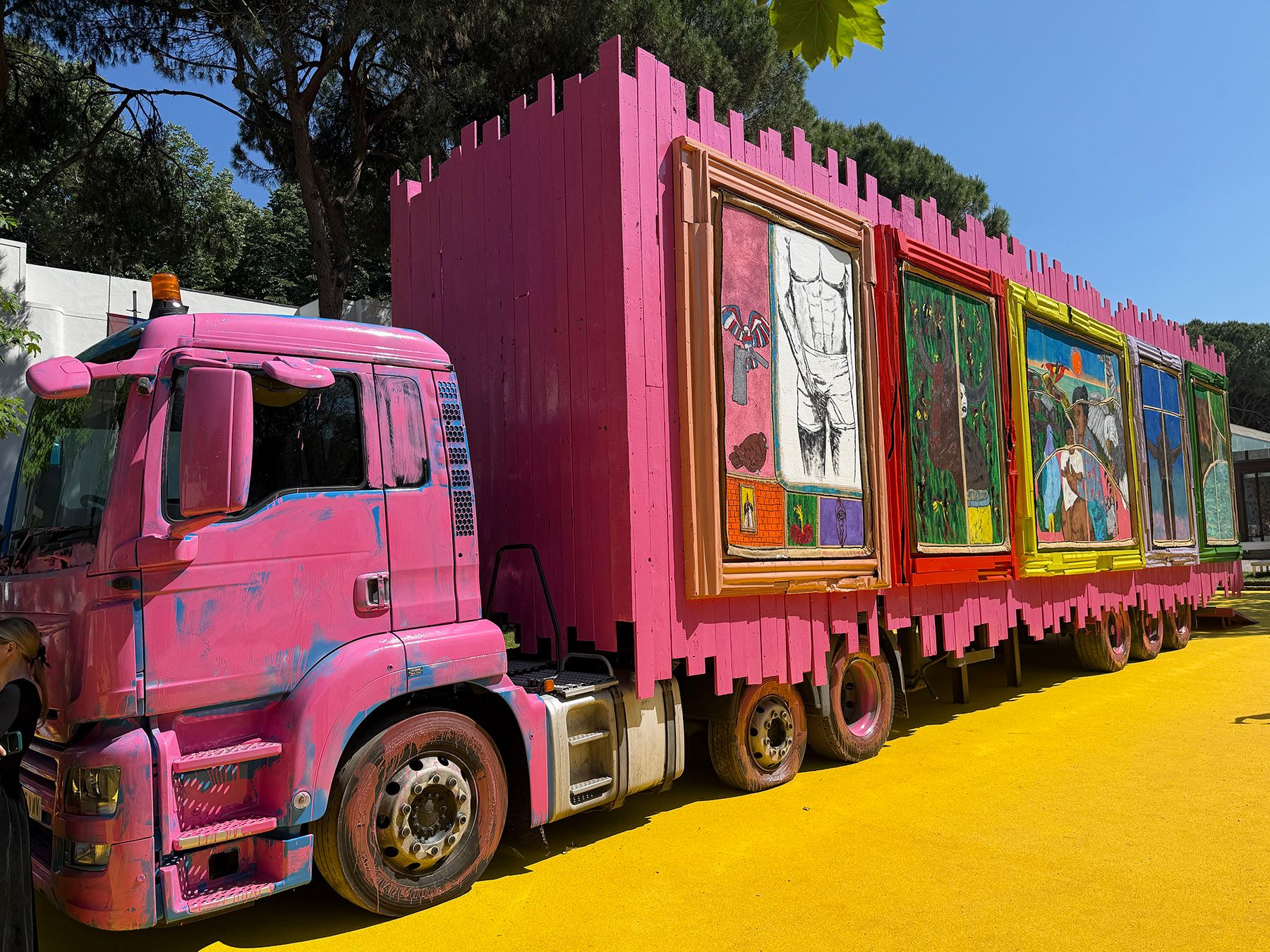 A large pink truck and trailer, with the trailer styled like a castle wall displaying framed artworks, rests on vibrant yellow ground outdoors.