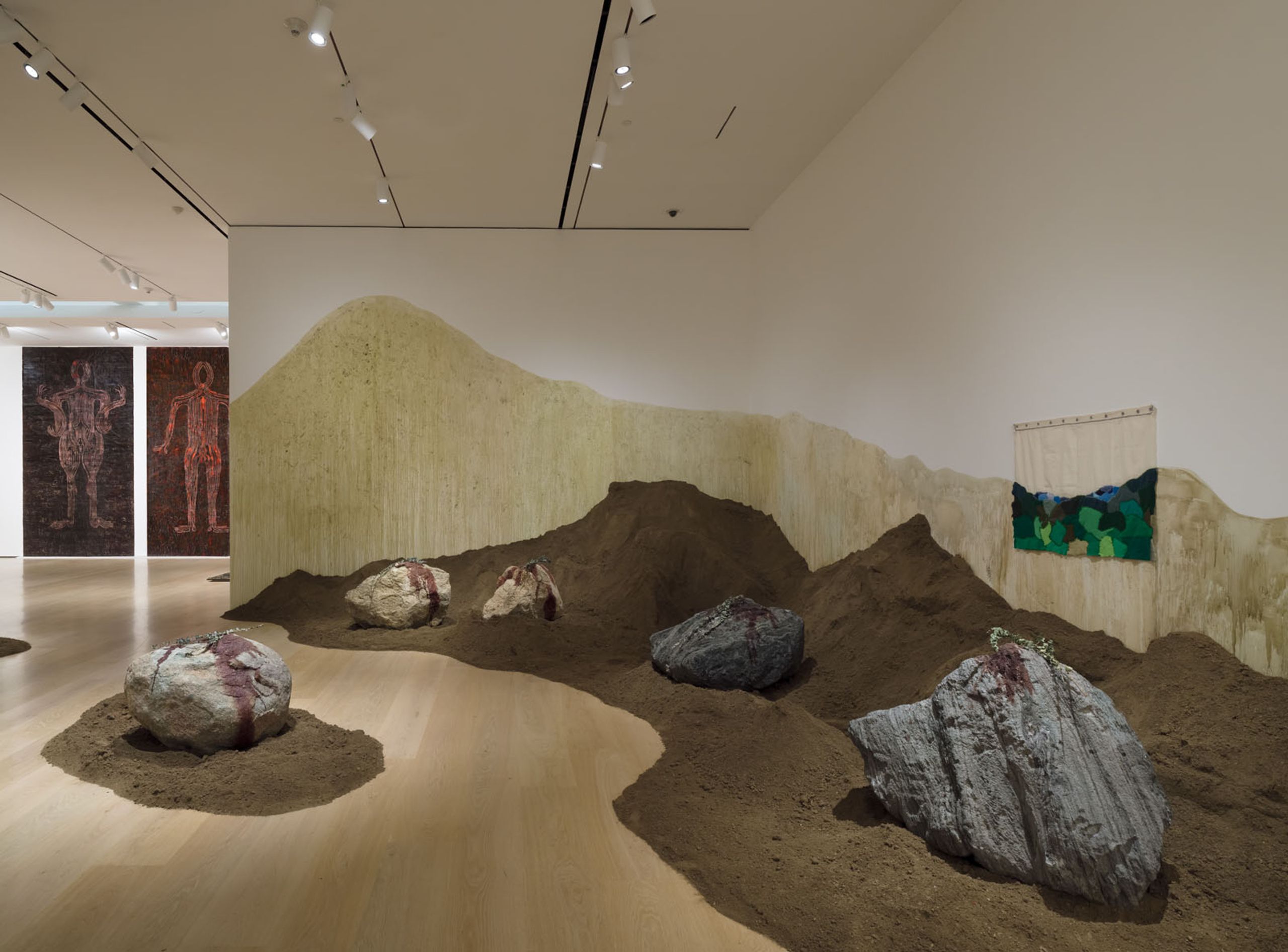 An art installation showing brown soil mounds and rocks with red markings on a light floor, backed by an undulating yellow-green textured wall. Two large artworks are visible in an adjacent room.