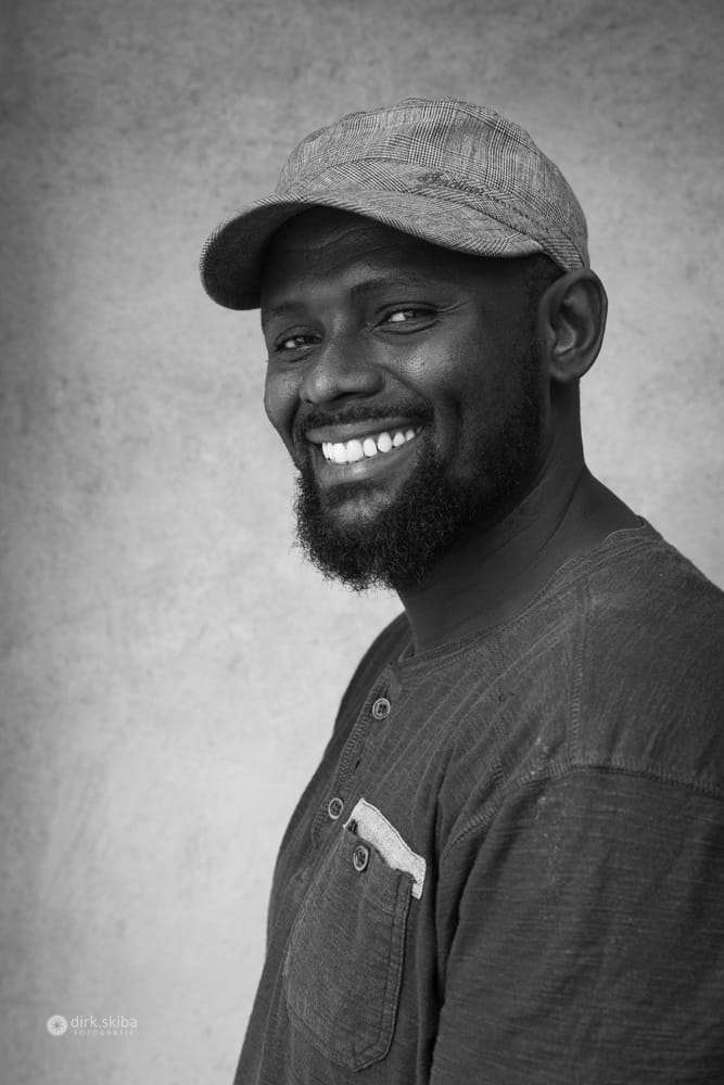 Black and white portrait of a broadly smiling man with a beard and a plaid cap.