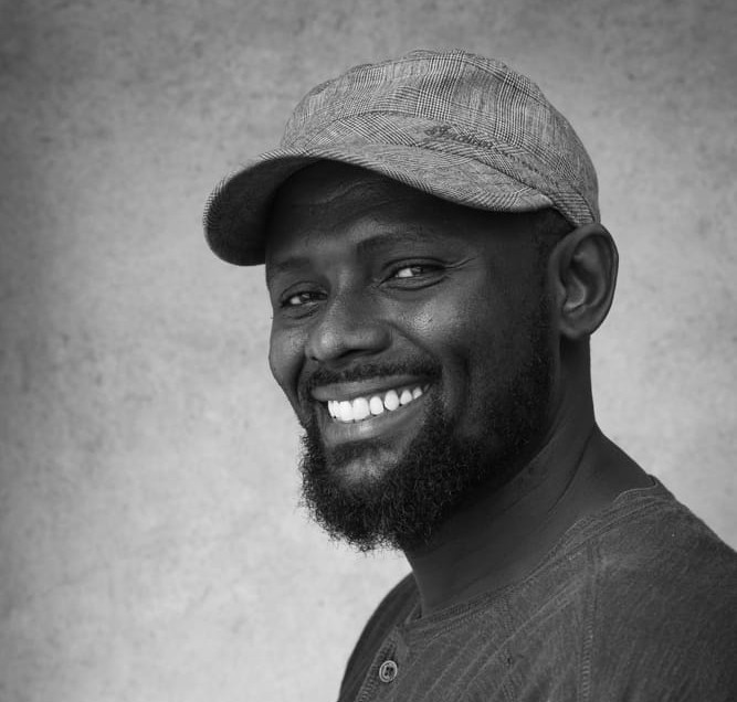Black and white portrait of a broadly smiling man with a beard and a plaid cap.