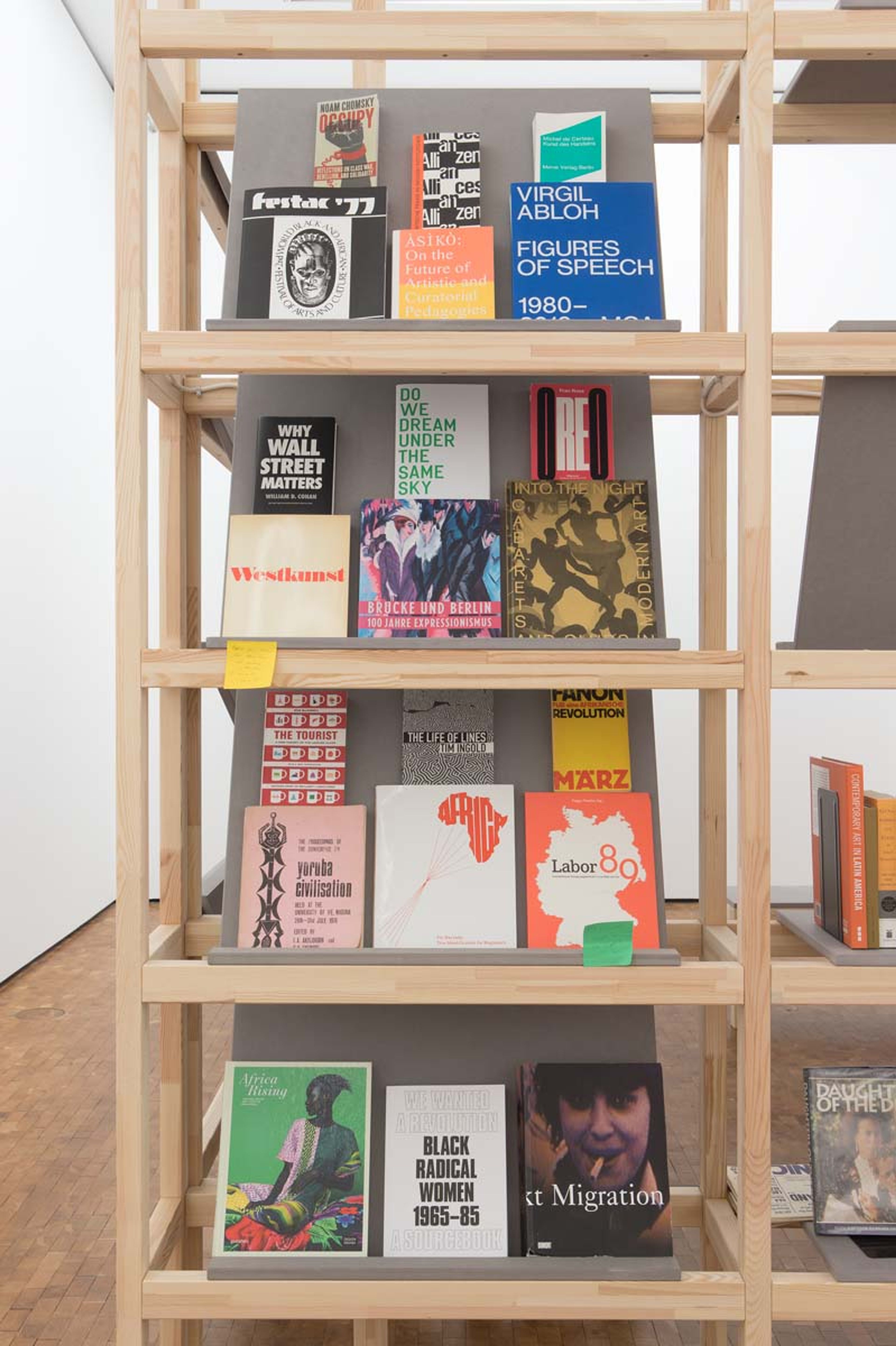 A light wood bookshelf displays a diverse collection of books and magazines, including titles like "Virgil Abloh," "Occupy," and "Black Radical Women."