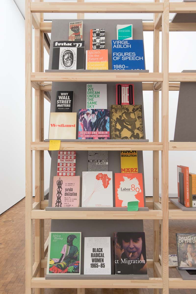 A light wood bookshelf displays a diverse collection of books and magazines, including titles like "Virgil Abloh," "Occupy," and "Black Radical Women."