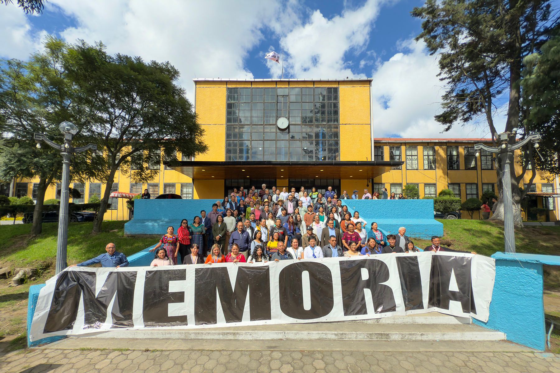 A large group of people stand in front of a yellow building, holding a banner that reads "MEMORIA".