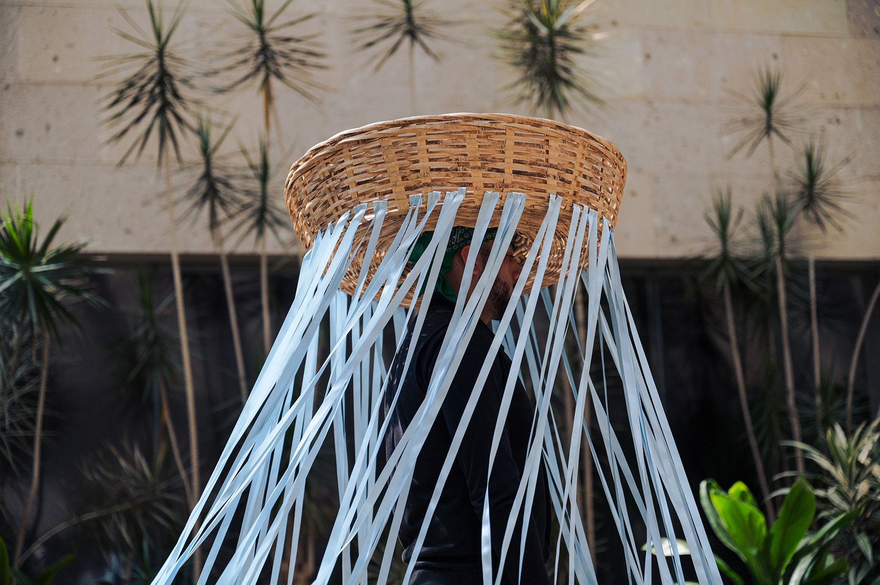 Person wearing a large woven basket on their head with light blue ribbons hanging down, obscuring their face.
