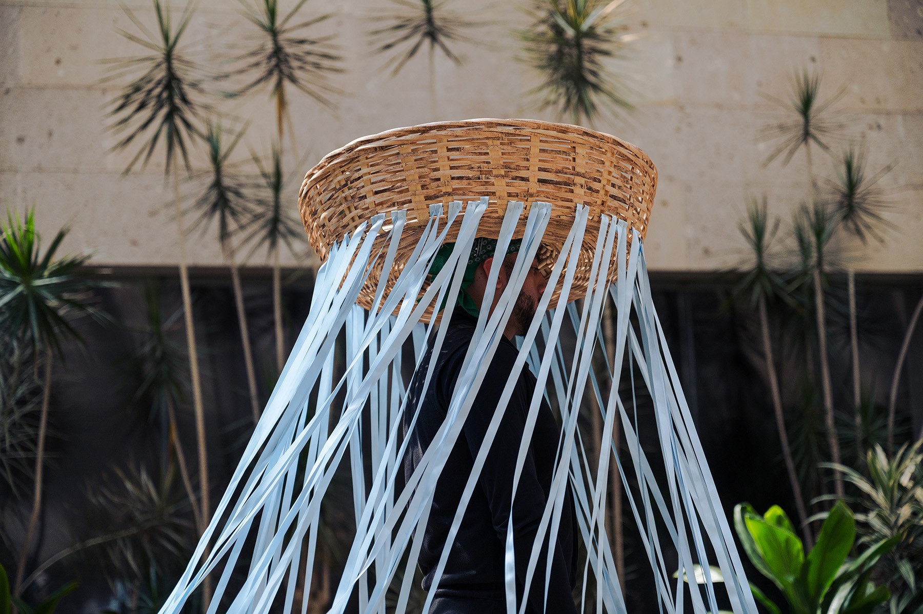 Person wearing a large woven basket on their head with light blue ribbons hanging down, obscuring their face.
