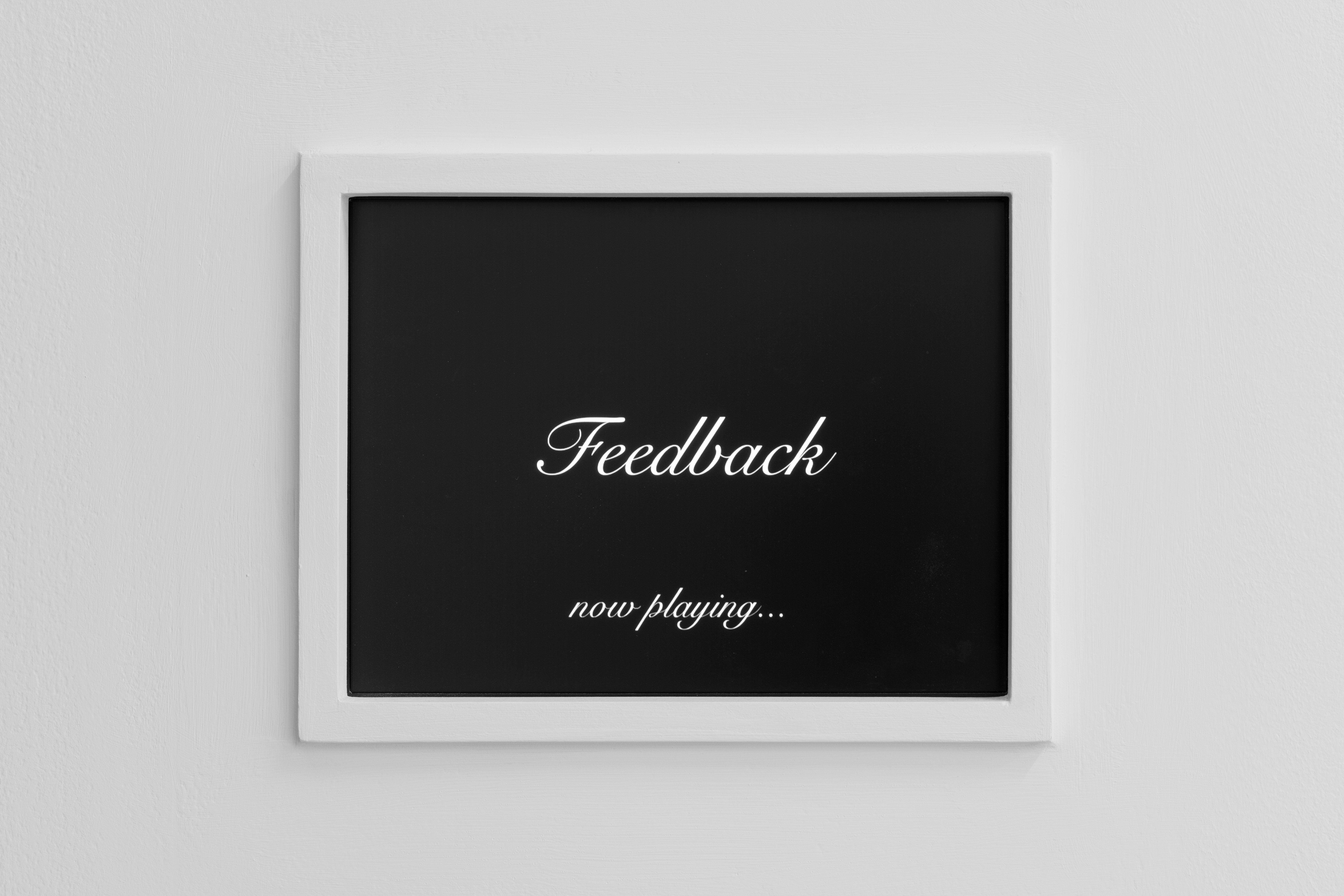 White framed picture on a white wall displaying "Feedback now playing..." on a black background.