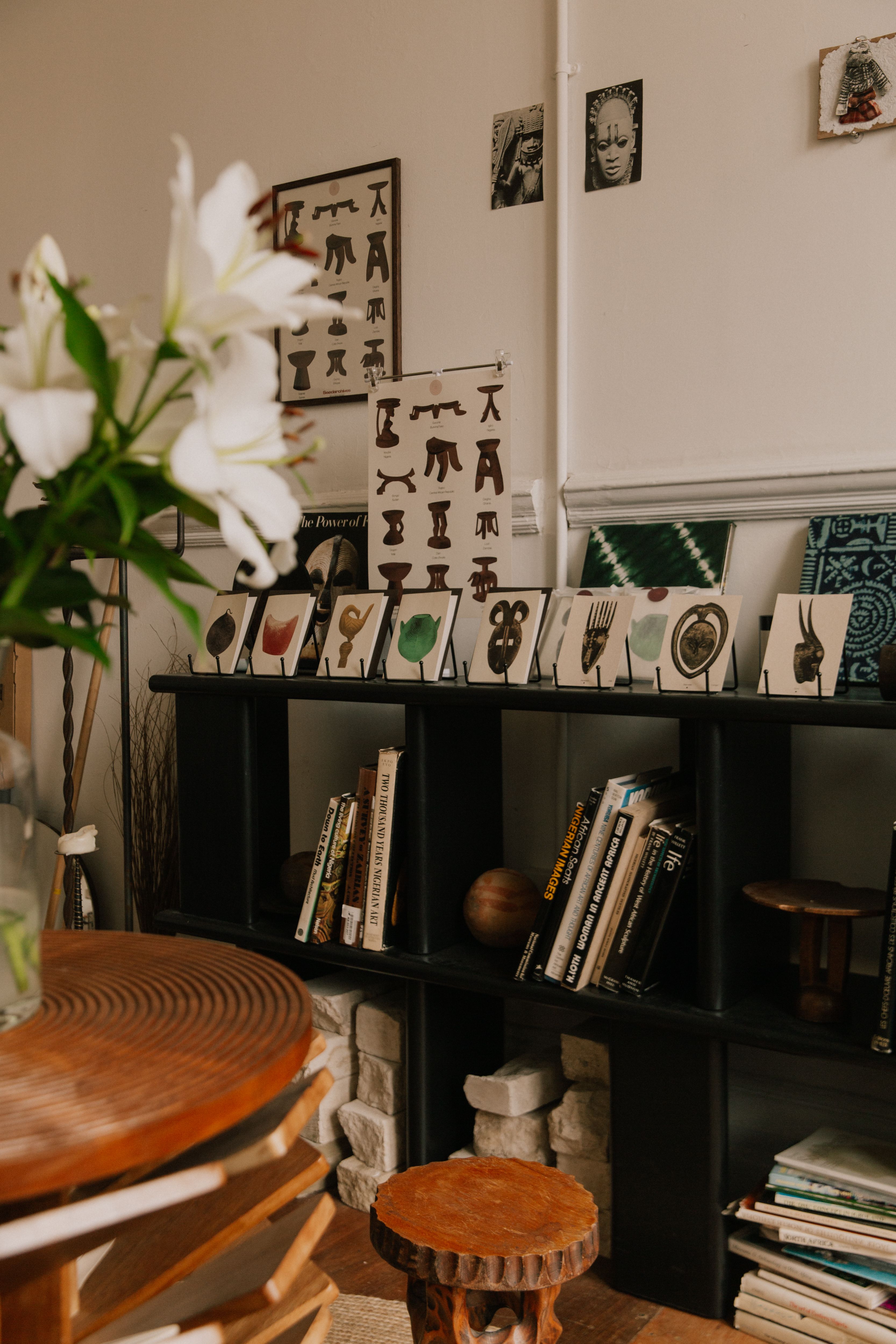 A room with a black bookshelf displaying books and African art, a round wooden table, and white lilies.