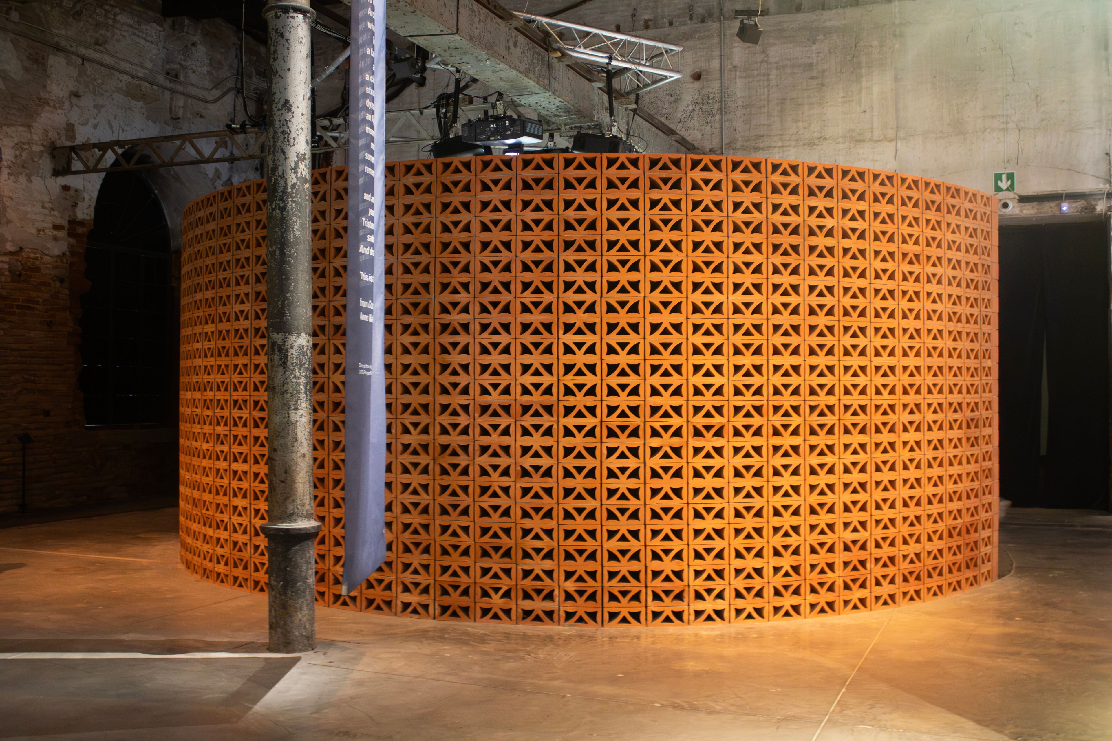 Curved wall of orange terracotta bricks with triangular perforations in an industrial building, with a tall blue banner.