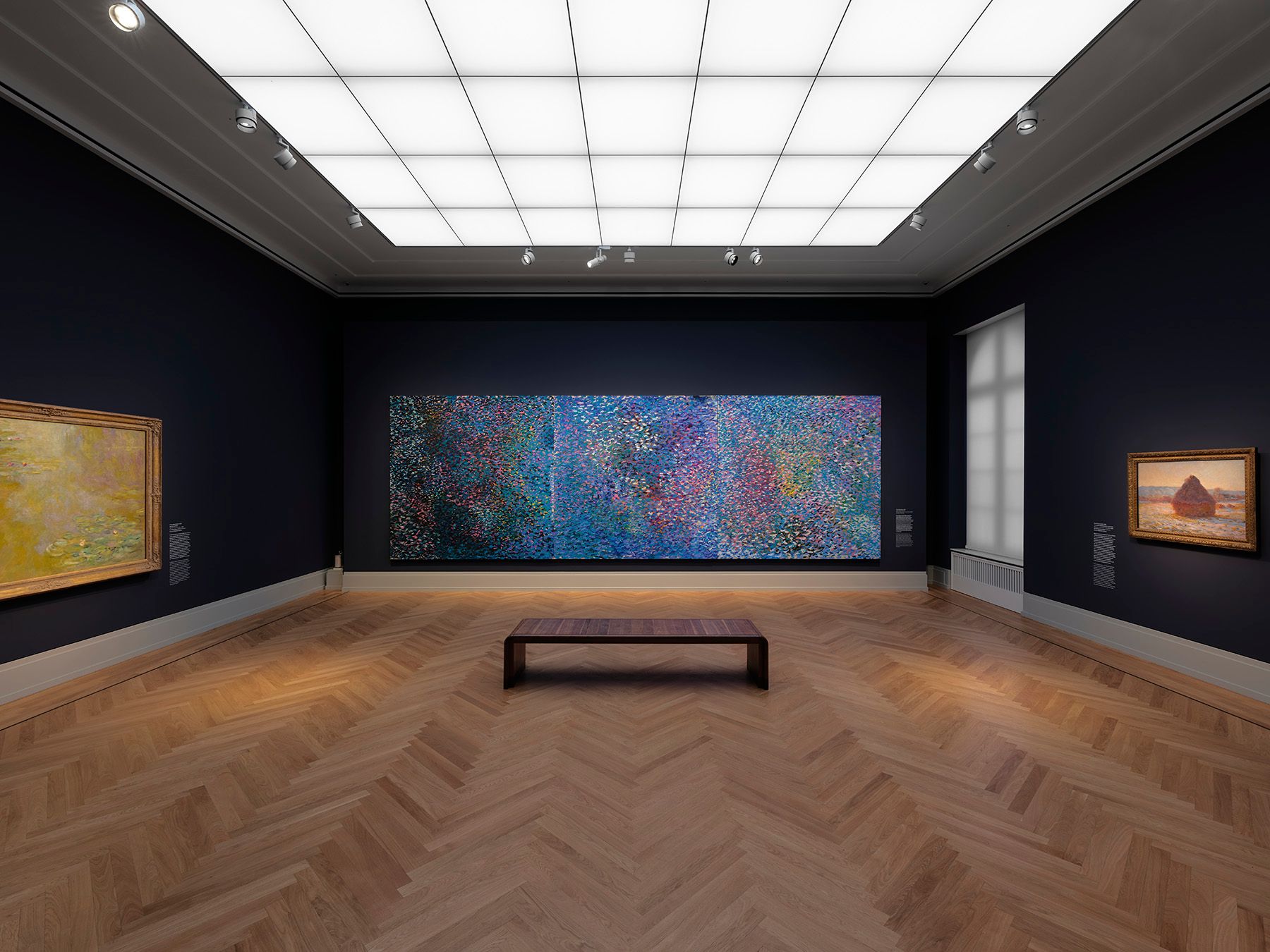 An art gallery with dark blue walls, wood floors, a long bench, and three framed paintings, including a large colorful multi-panel artwork.