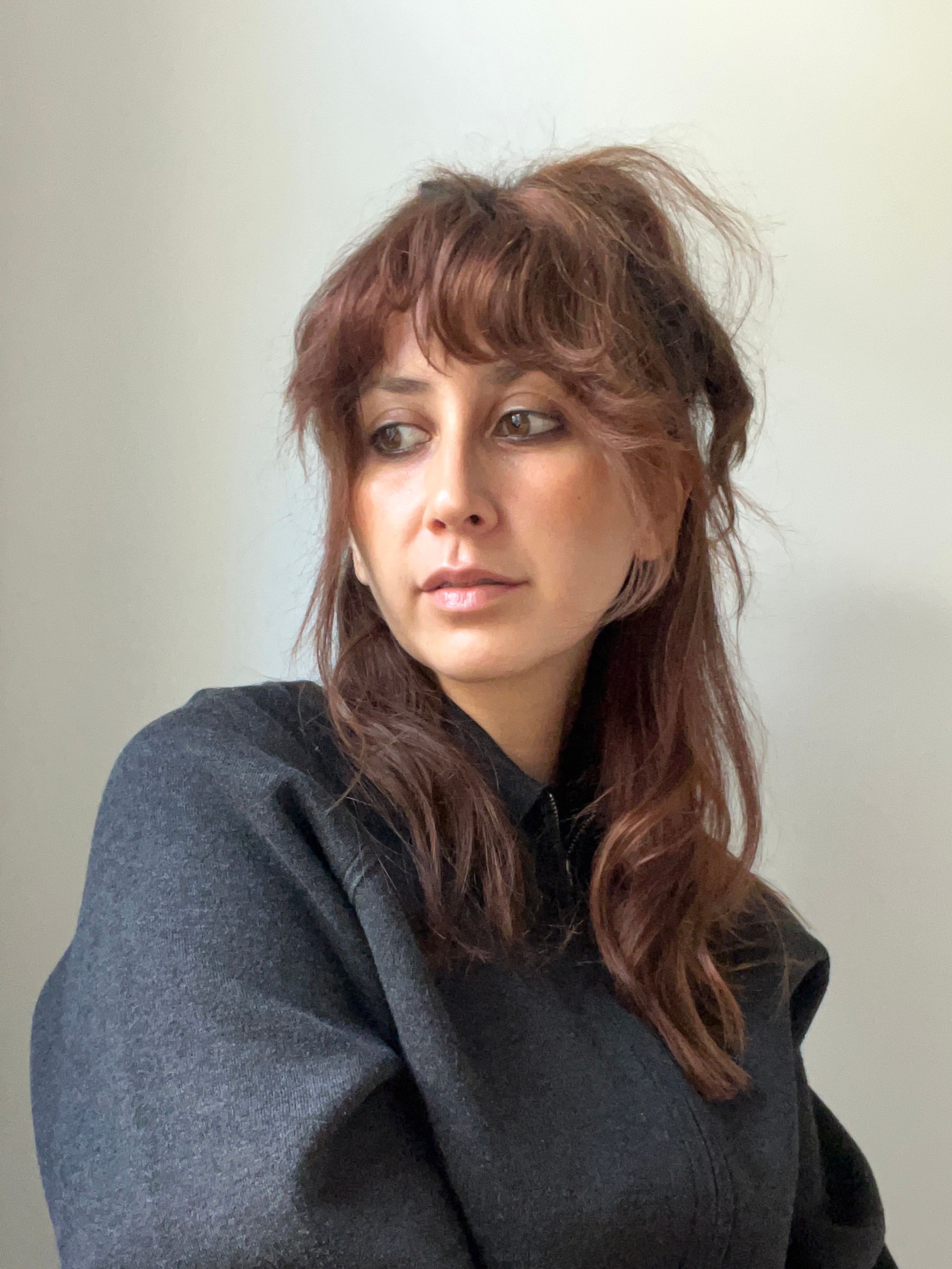 A woman with reddish-brown hair and bangs, wearing a dark grey jacket, looks left.