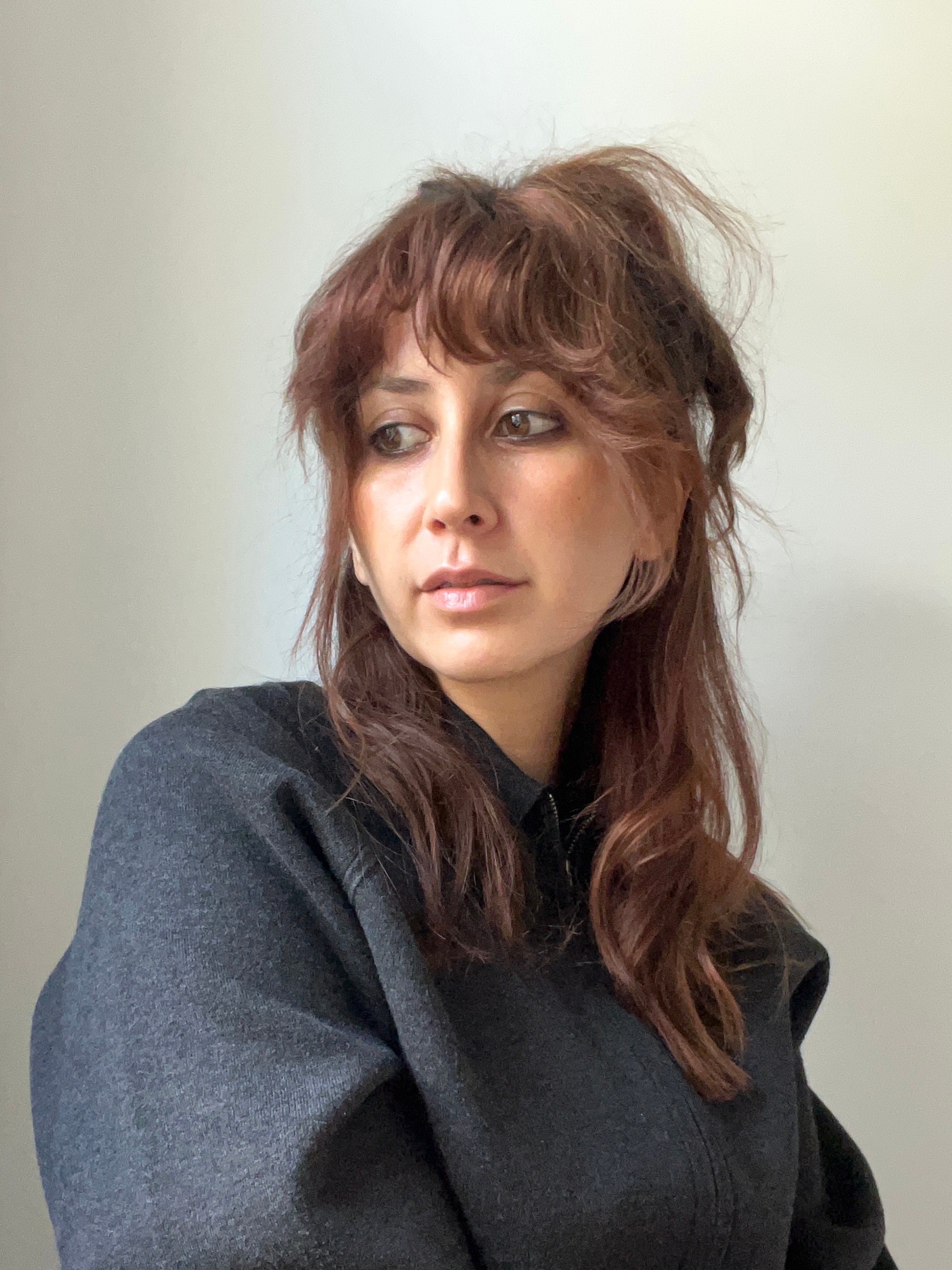 A woman with reddish-brown hair and bangs, wearing a dark grey jacket, looks left.