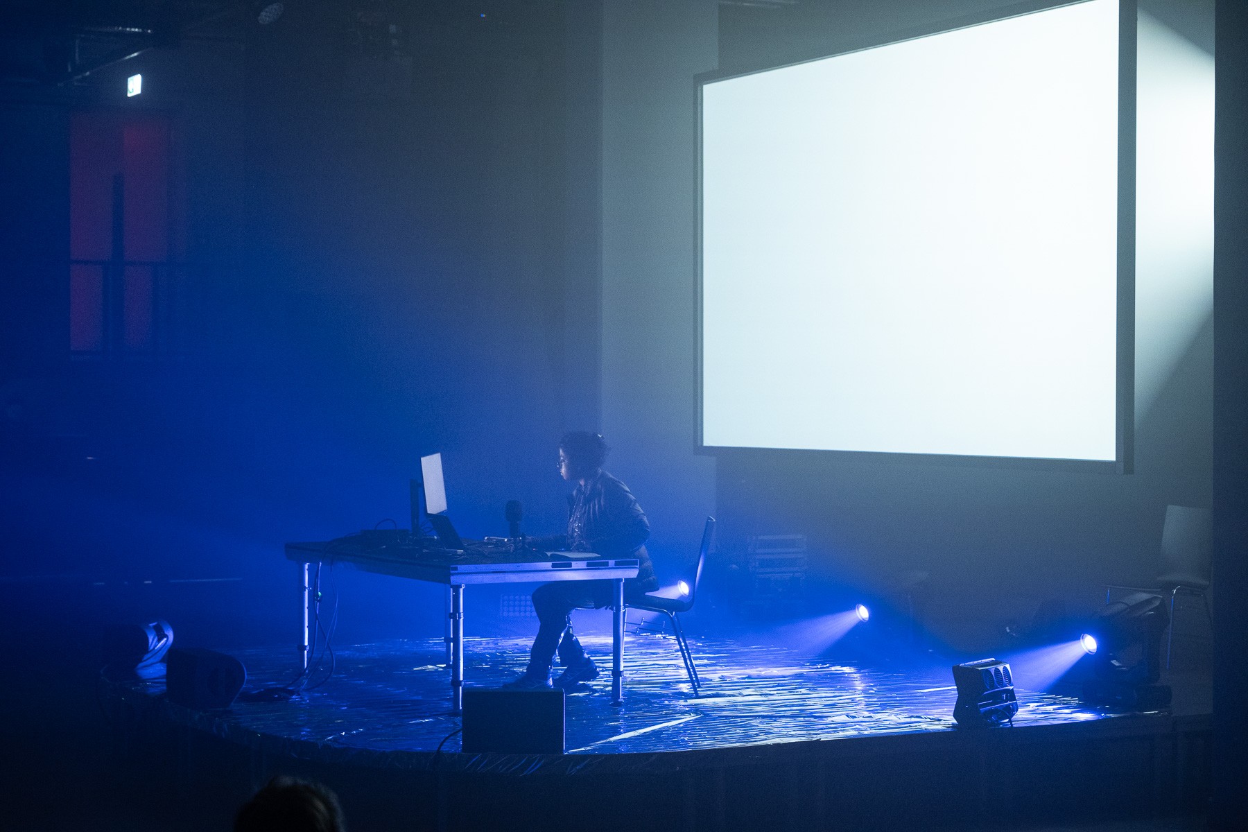A person works at a table with a laptop and sound equipment on a blue-lit stage, with a large blank screen behind them.