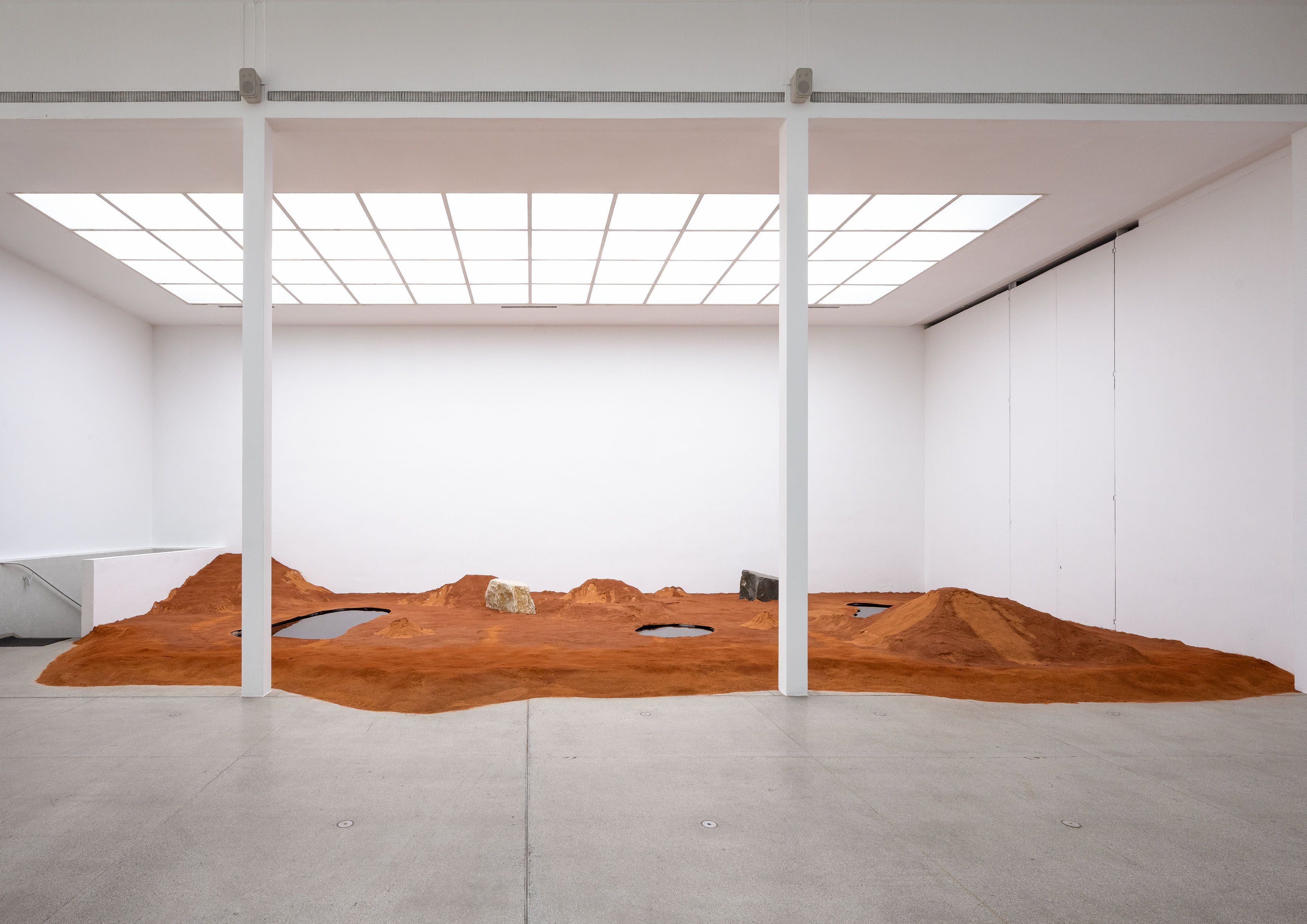 Reddish-brown sculpted desert landscape with dark pools and a rock in a white gallery.