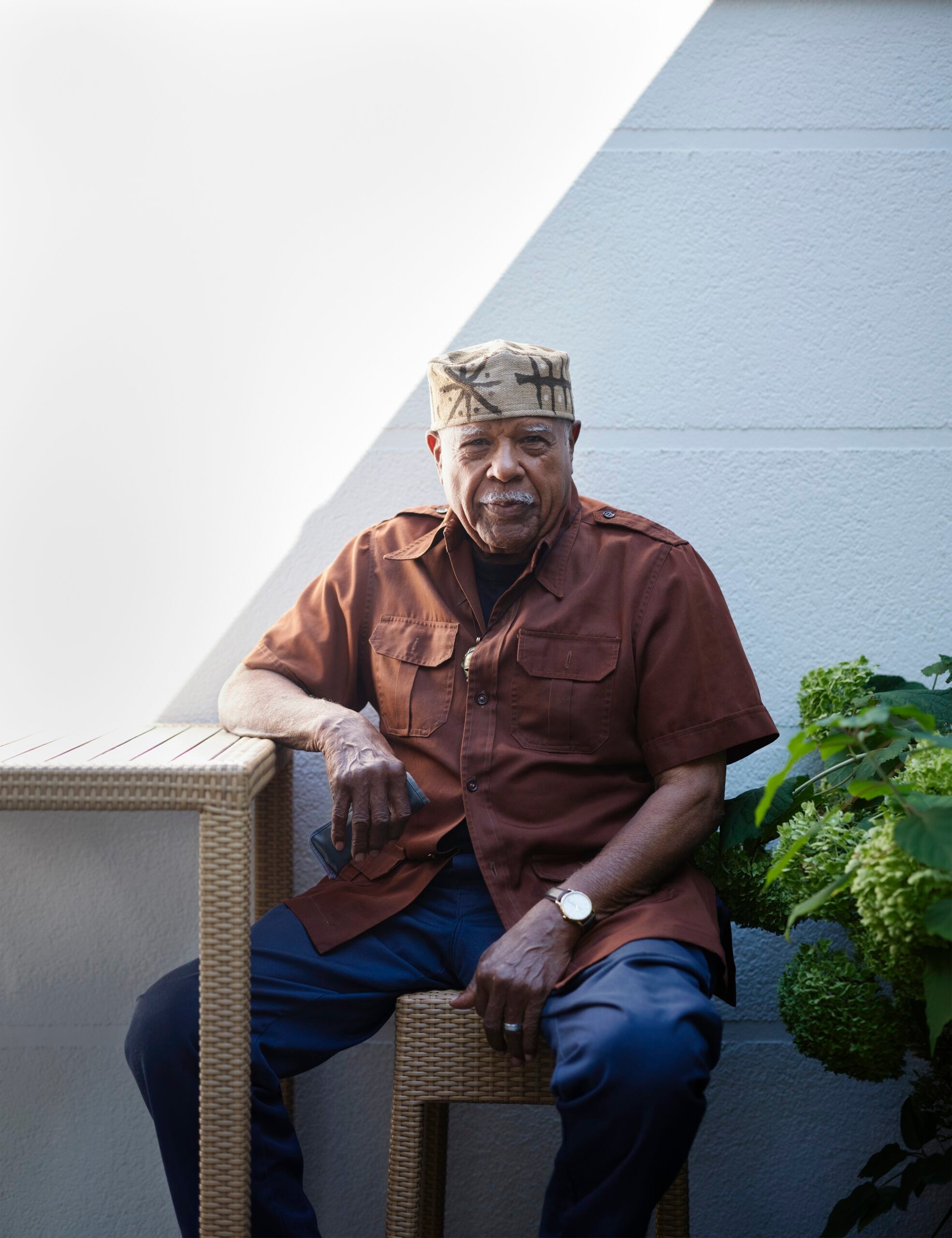An older Black man with a mustache sits on a wicker chair, wearing a brown shirt and patterned cap, looking at the camera against a white wall with a diagonal shadow.