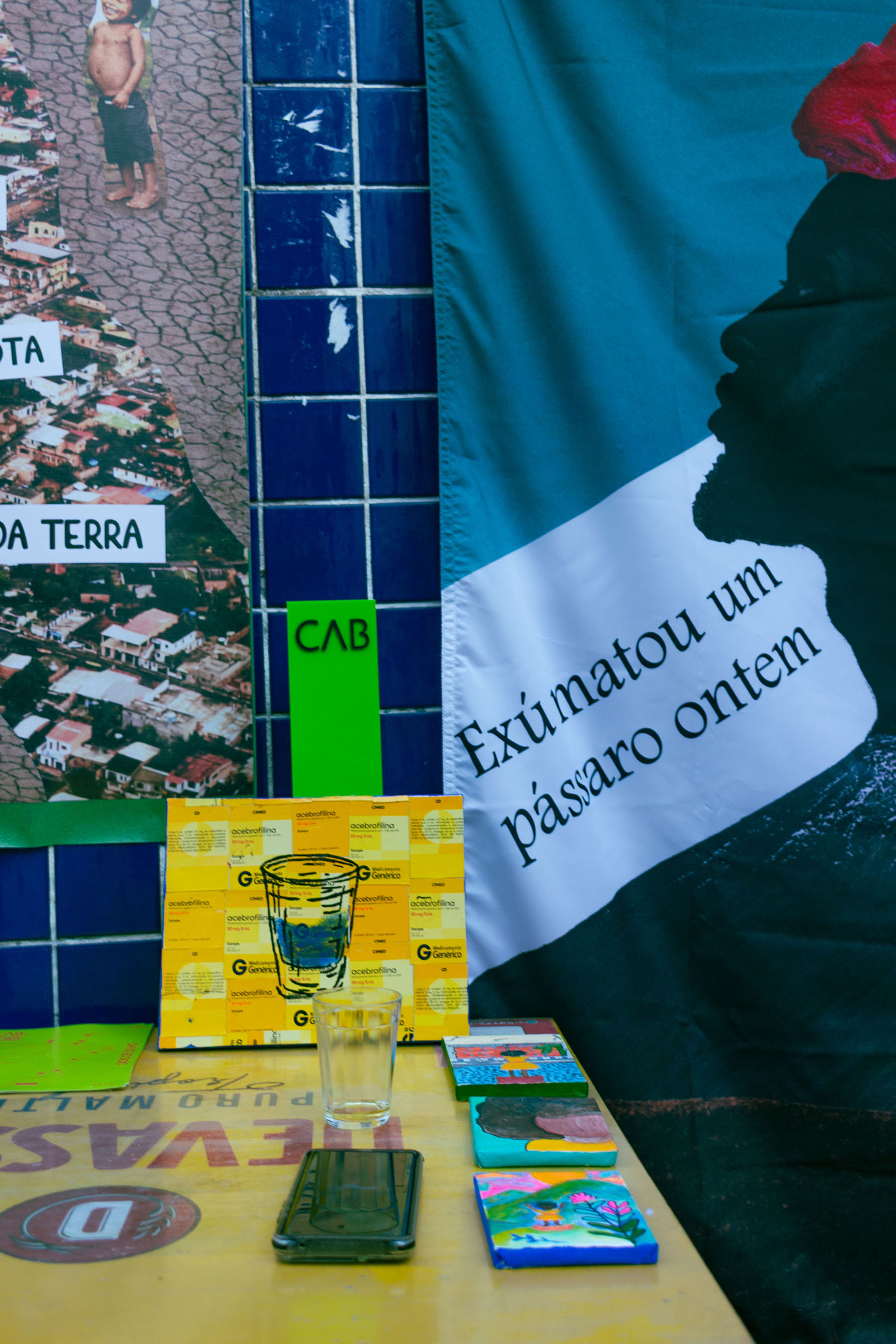 A wall with a photo collage, blue tiles, and a teal banner featuring a silhouette and Portuguese text, above a yellow table with a glass, a phone, and small colorful painted blocks.